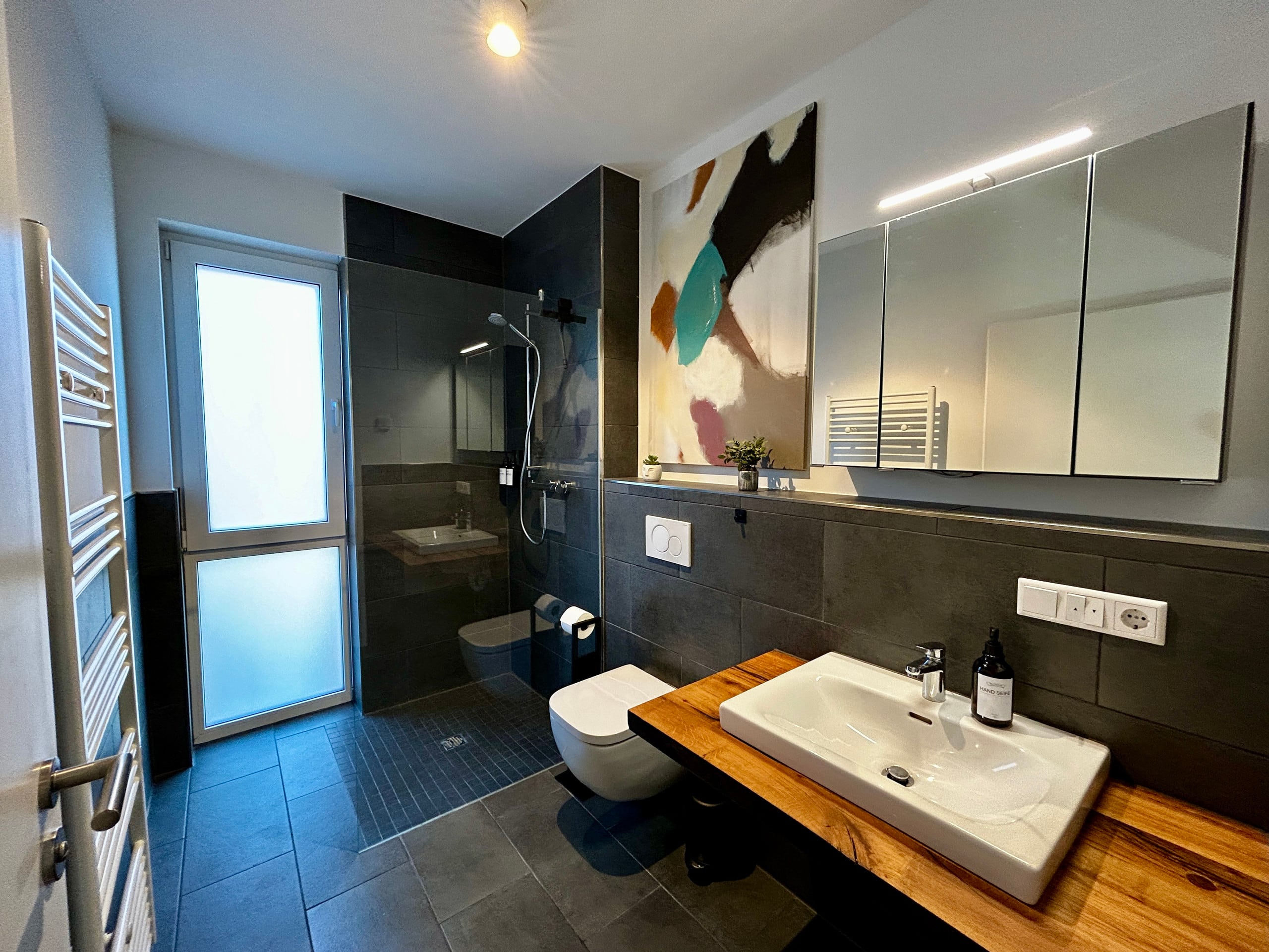Modern bathroom with grey tiles, a glass-partitioned walk-in shower, wall-hung toilet and a white basin on a warm wooden countertop beneath a lit mirror.