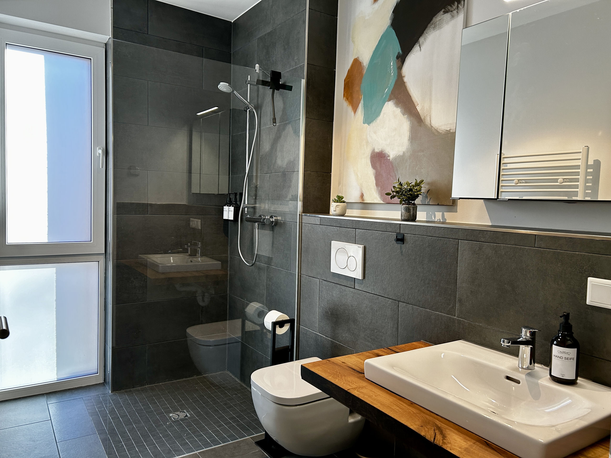 Closer view of the same bathroom, focusing on the rainfall shower with black fittings behind the glass panel, beside the wall-hung toilet and wooden vanity.