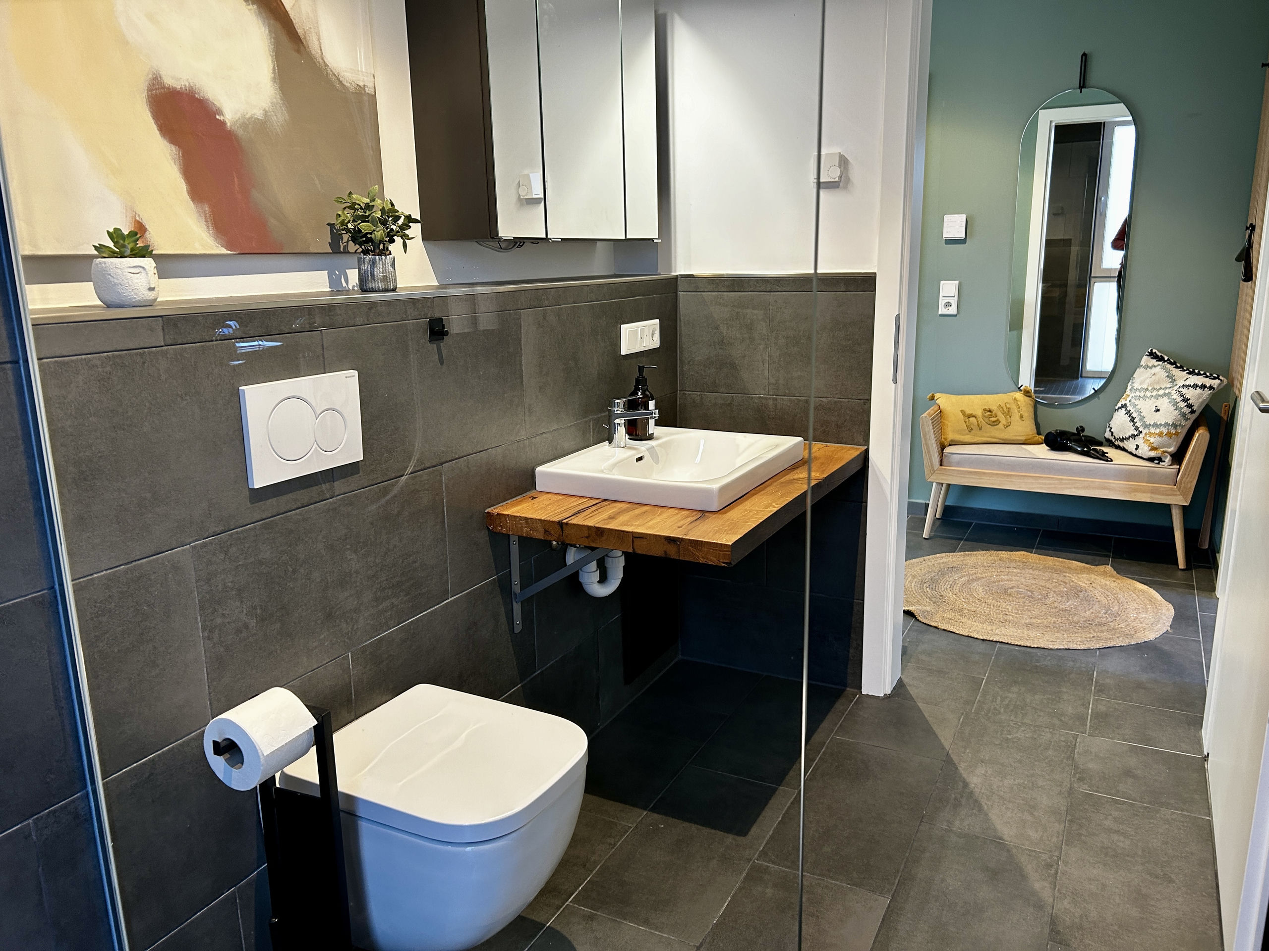 Wall-mounted wooden vanity with a white basin and wall-hung toilet in the grey-tiled bathroom; through the doorway a green hall with a bench and jute rug is visible.