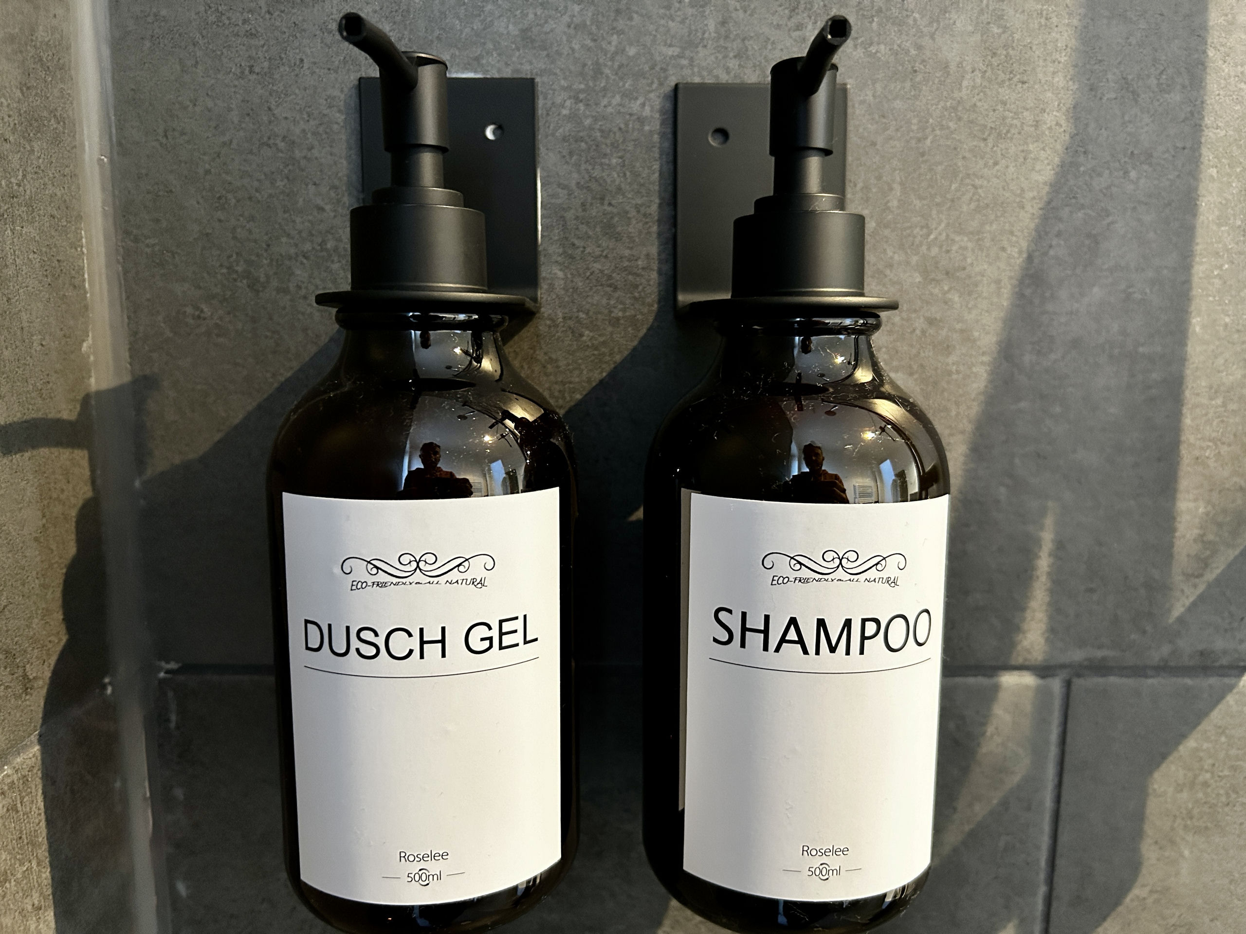 Close-up of two dark refillable pump bottles mounted on a grey tile wall, labelled "Dusch Gel" and "Shampoo" on white labels.