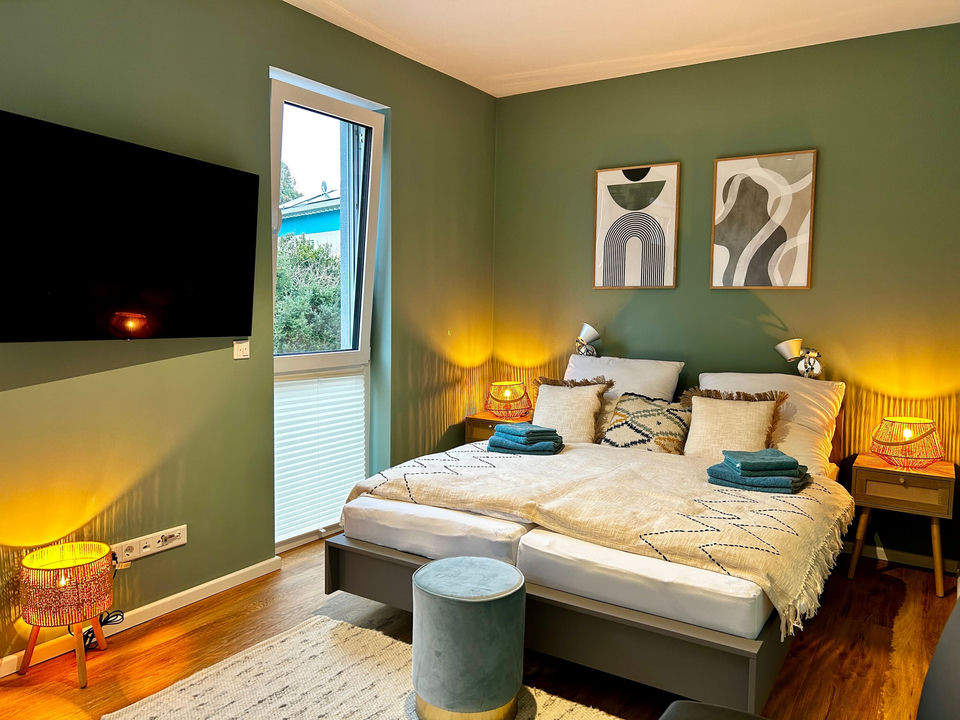 Bedroom with sage-green walls, a double bed dressed in a cream zigzag throw and cushions, two framed abstract prints, a wall-mounted TV and warm woven lamps.