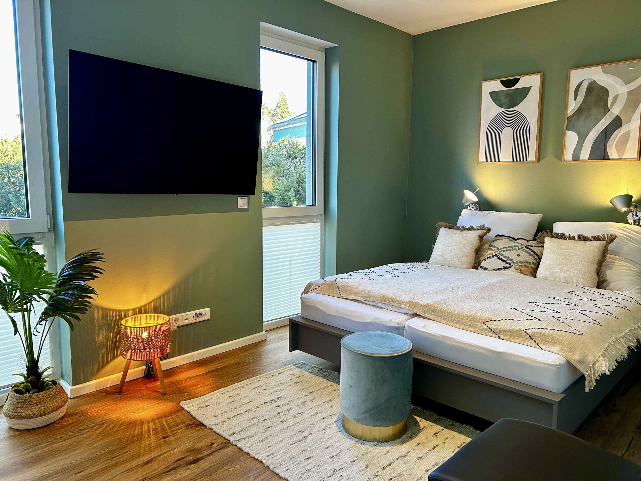 Same bedroom seen from the foot corner: flat-screen TV between two tall windows, potted palm in a basket, teal velvet stool and rug on the wooden floor.