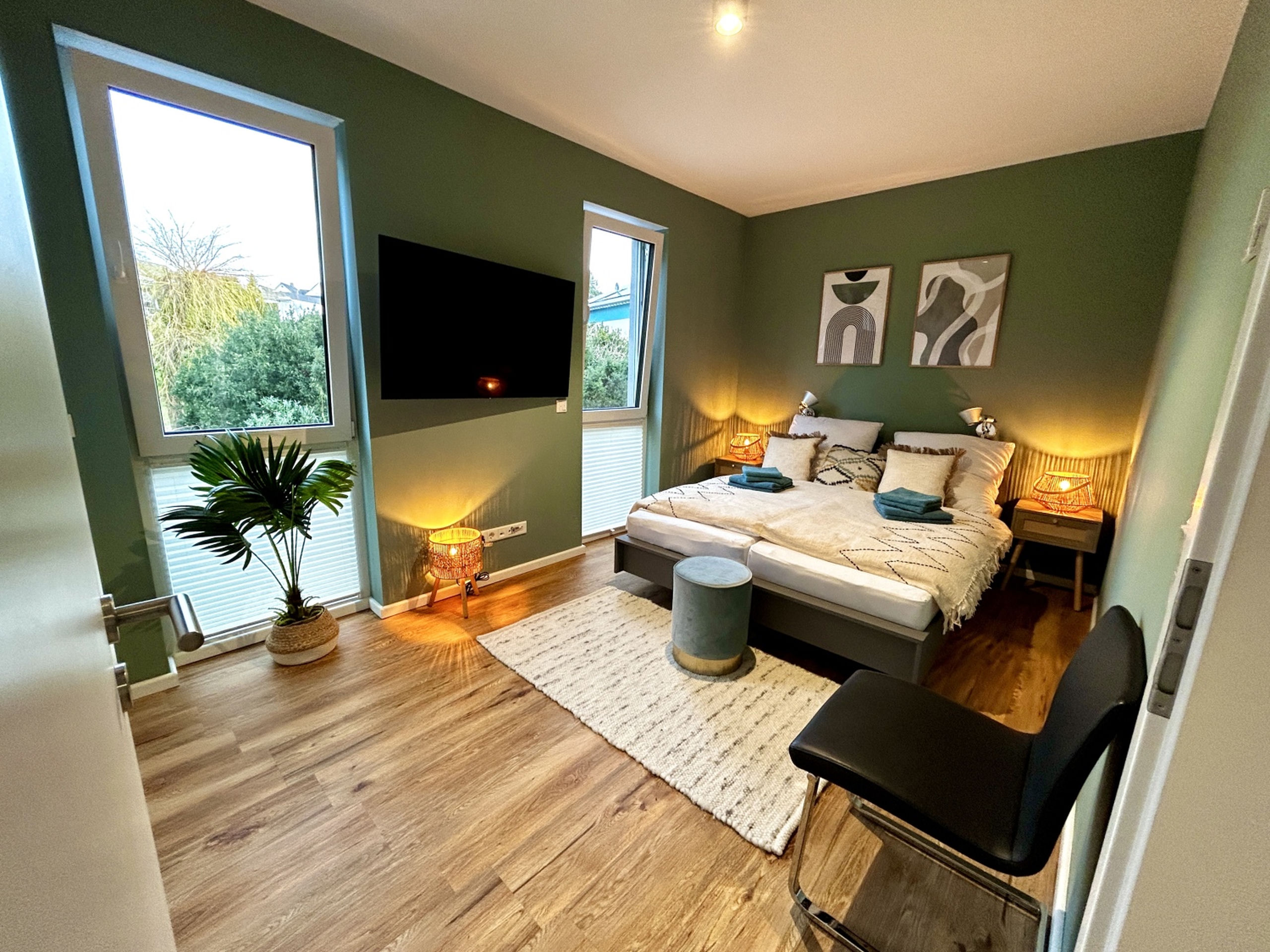Wide view of the green-walled bedroom from the doorway, showing the wall TV between two tall windows, the bed beyond, a palm plant, woven lamp and a black chair in the foreground.