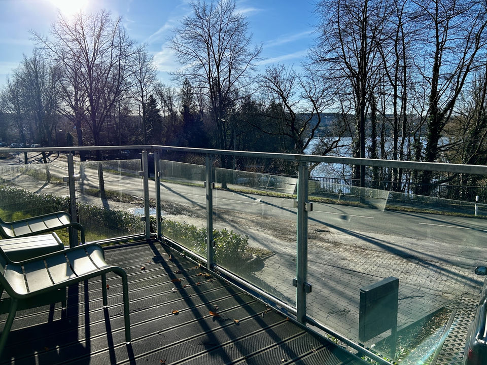 Balcony with glass railing and two metal chairs looks over a quiet road and bare winter trees towards a stretch of water under a bright blue sky.