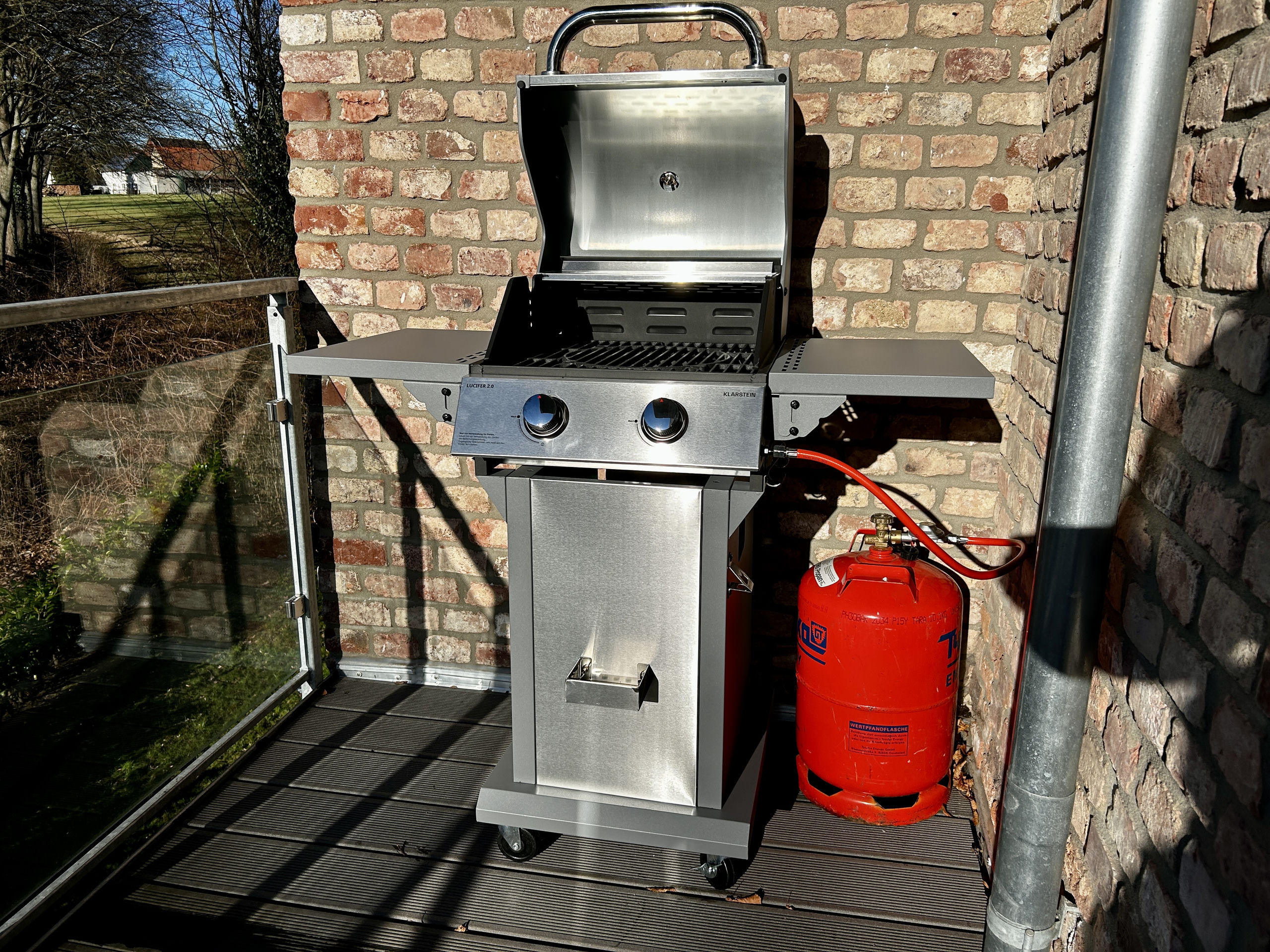 Stainless steel gas grill with its lid open stands on the wooden balcony deck against a brick wall, beside a red propane gas cylinder.