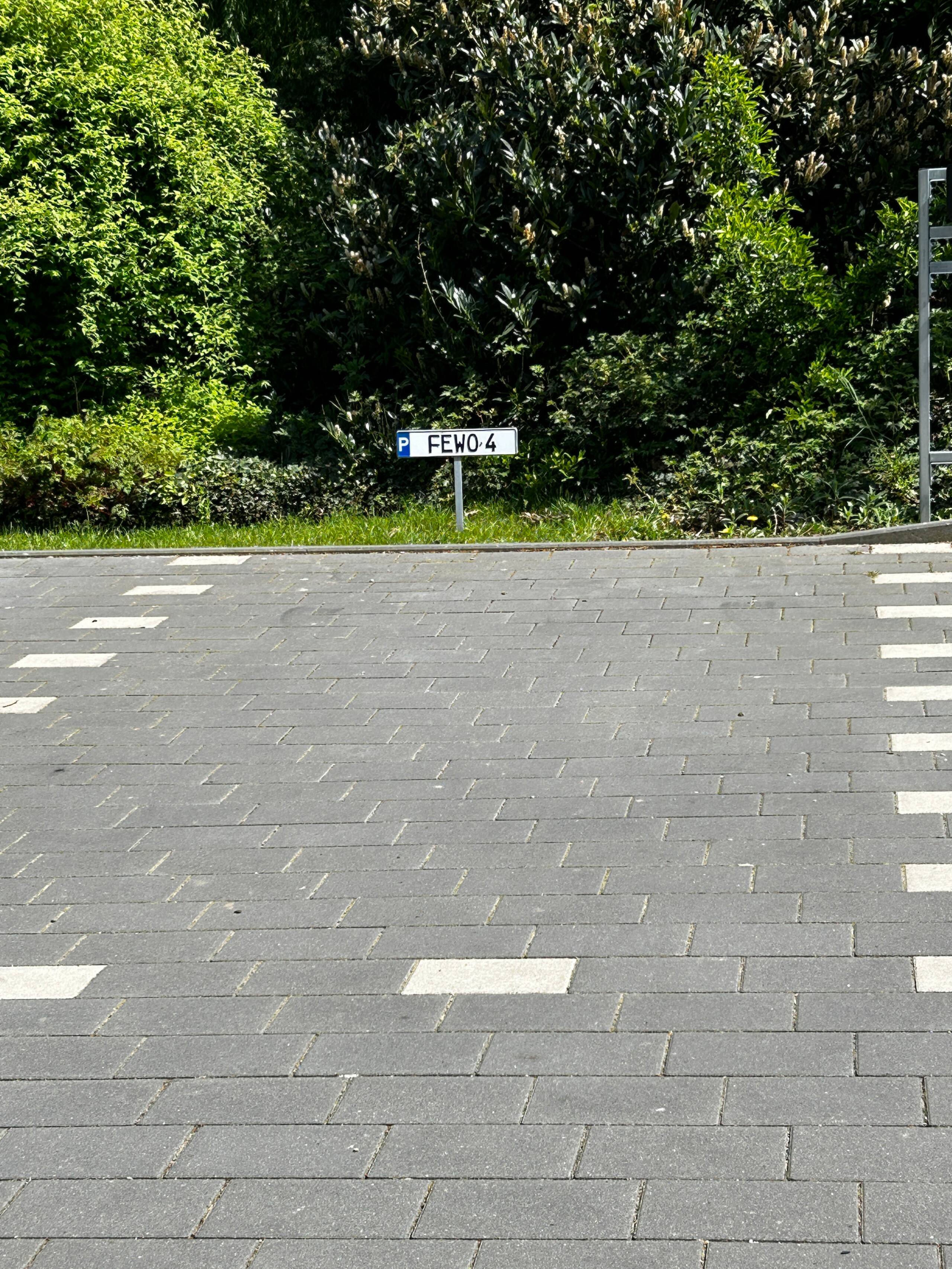 Paved parking area with a small white parking sign reading "P FEWO 4" at the edge of a leafy green hedge.