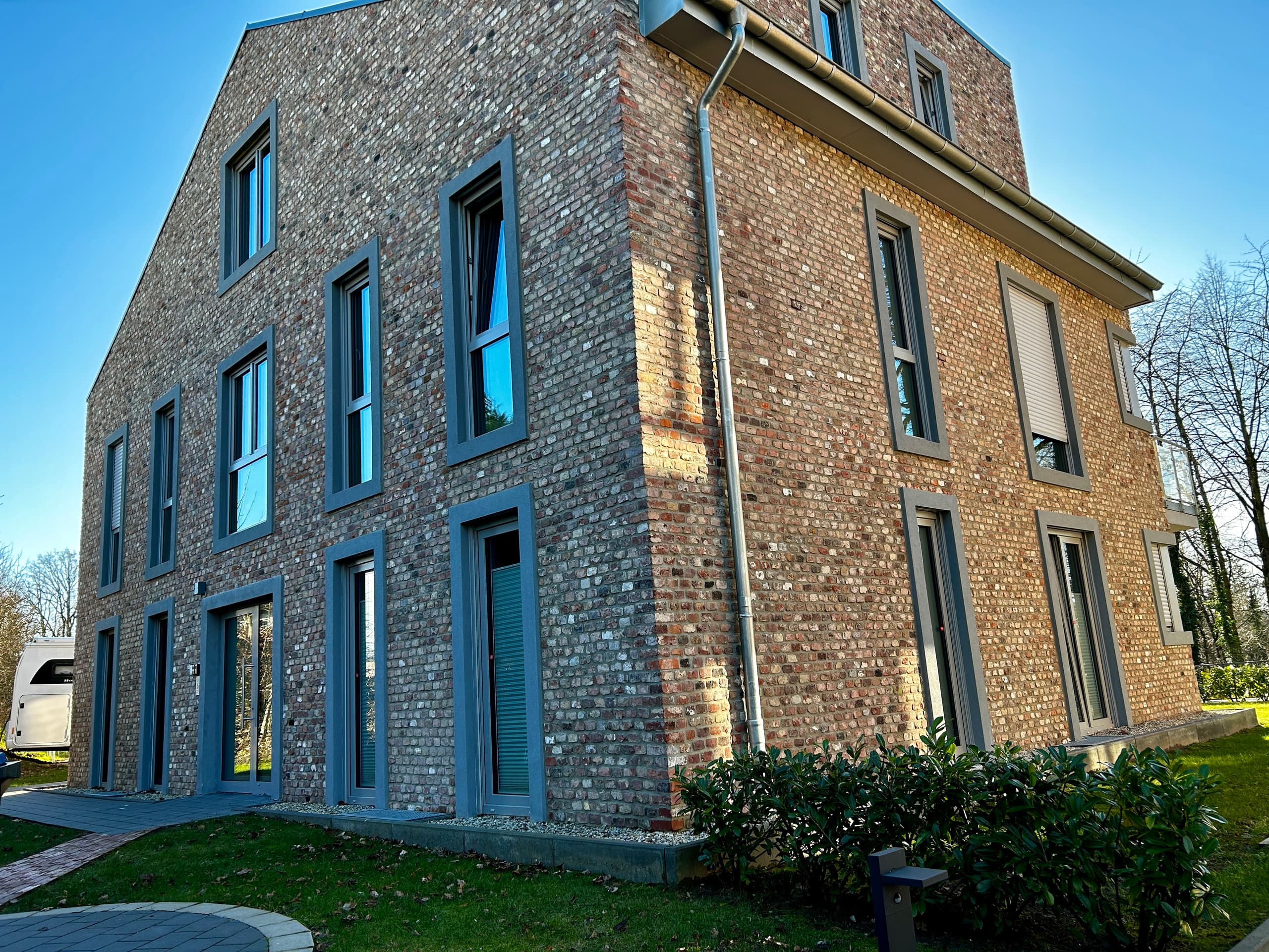 Corner view of the house's red brick facade with tall grey-framed windows and ground-floor doors, lawn and shrubs in front under a clear sky.