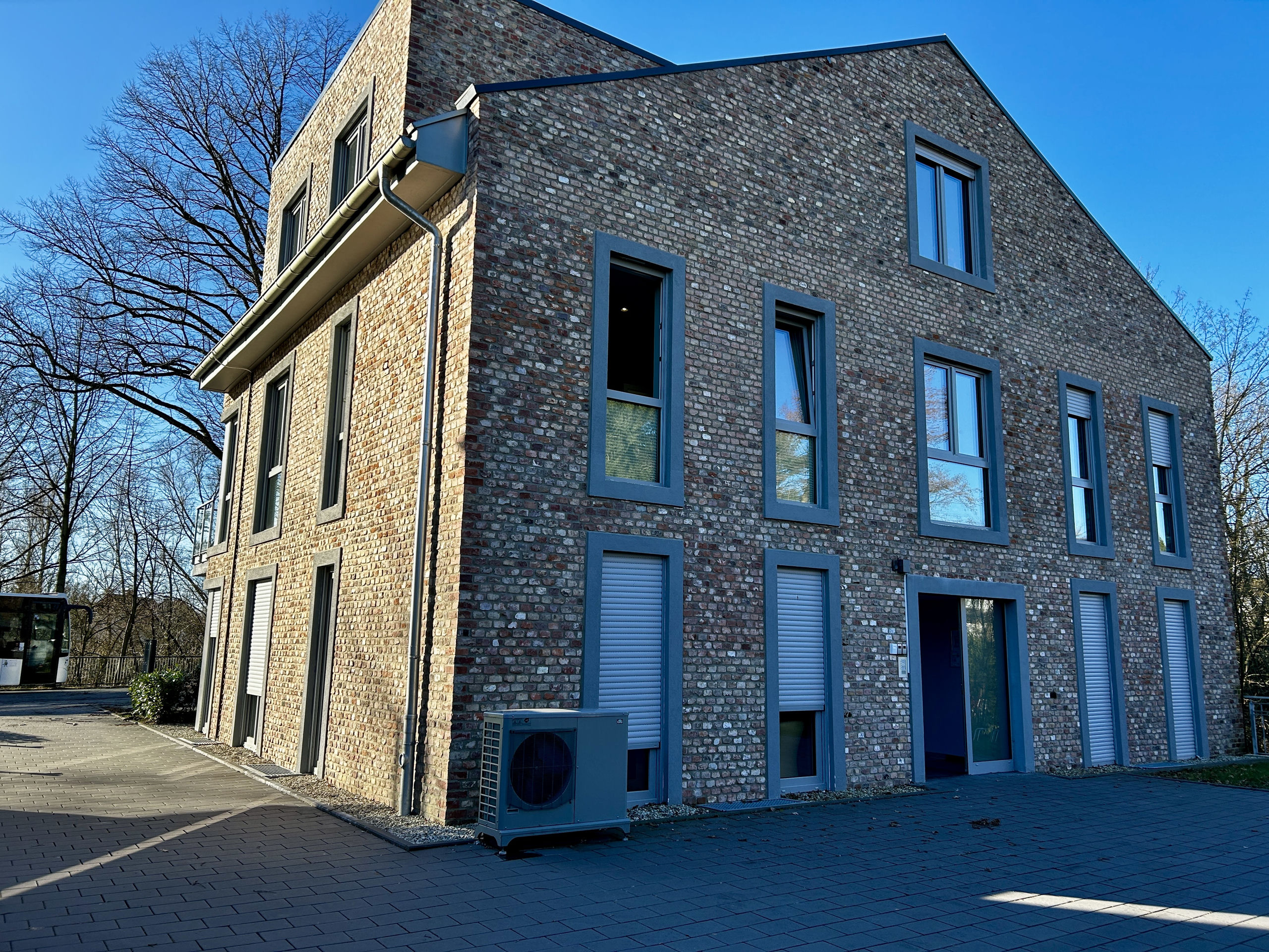 The same brick house seen from another corner, showing tall windows with grey shutters, a paved walkway and a heat pump unit at the wall.
