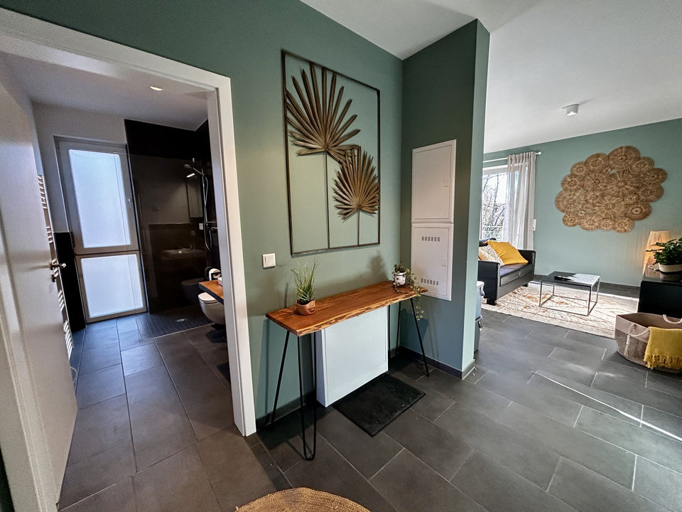 Hallway with grey tile floor and green walls, a slim wooden console table under a palm-motif mirror; the bathroom doorway is on the left and the living room opens up on the right.