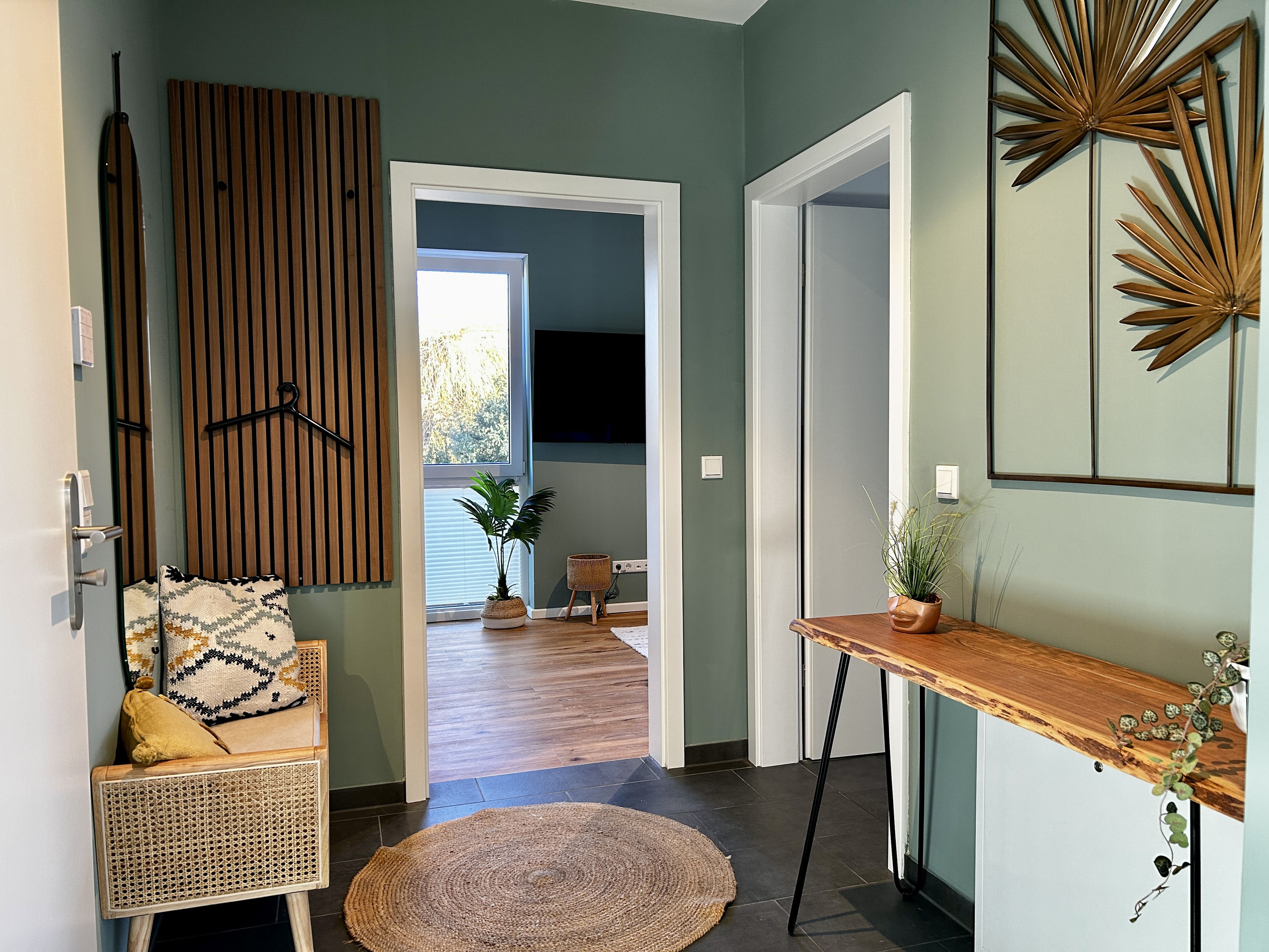 Entrance area with slatted wood coat panel, narrow mirror and rattan bench; through the open door the bedroom with its palm plant and wall TV is visible.