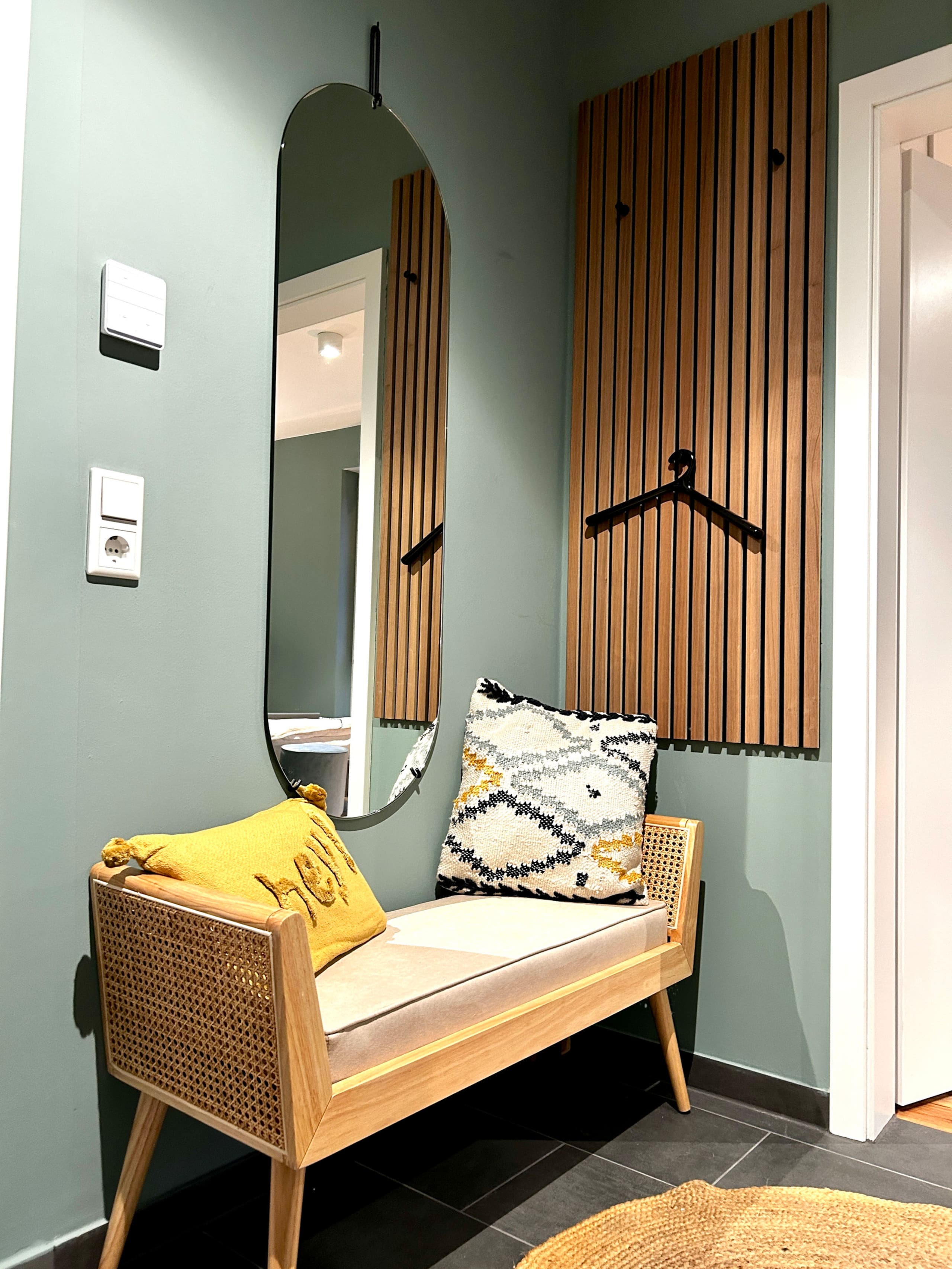 Vertical detail shot of the hallway bench with rattan sides, a yellow cushion and patterned pillow, beneath an oval hanging mirror and slatted coat panel.