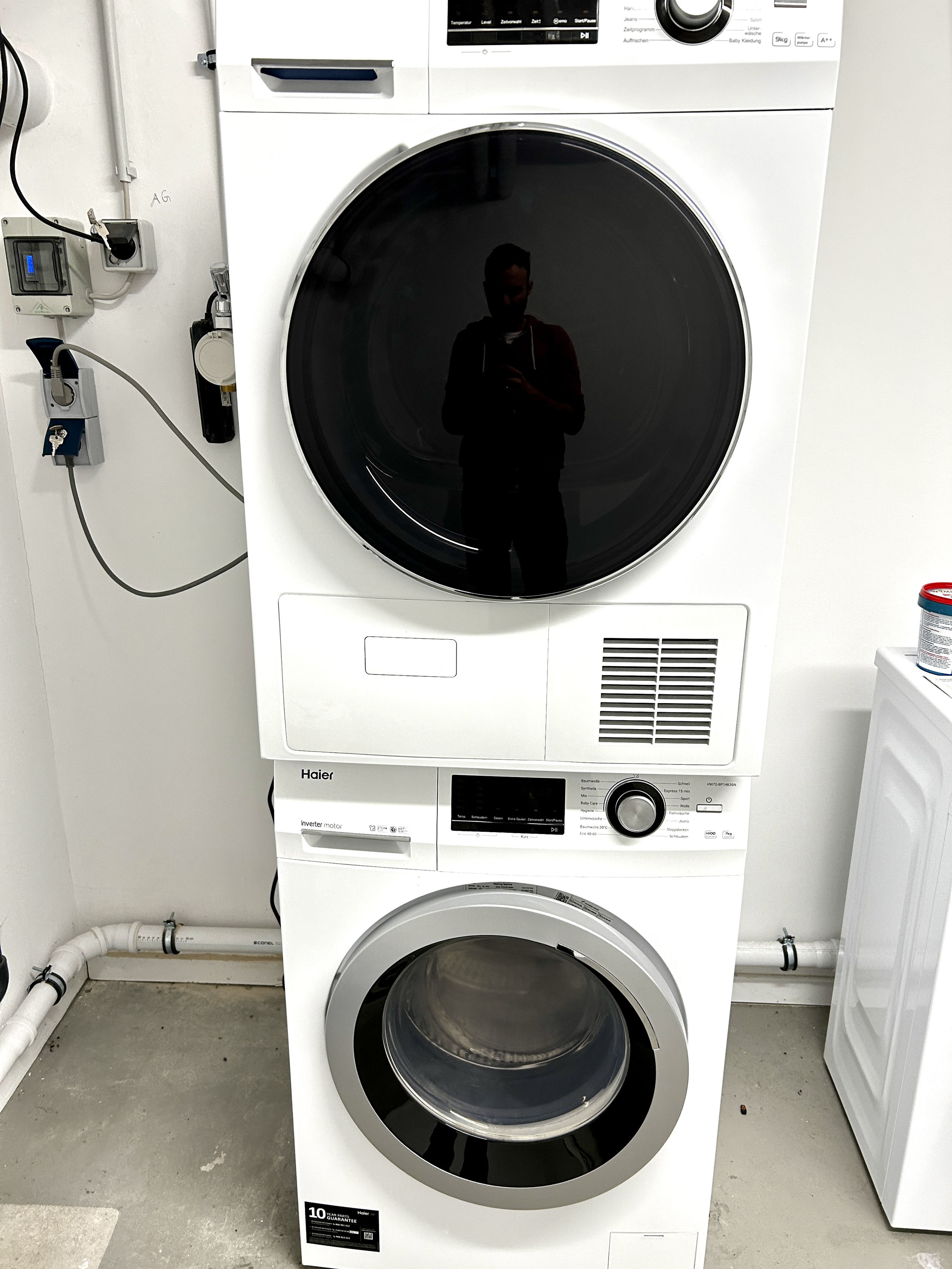 Vertical photo of a stacked washer and tumble dryer in a white utility room, with sockets and plumbing visible on the wall beside them.