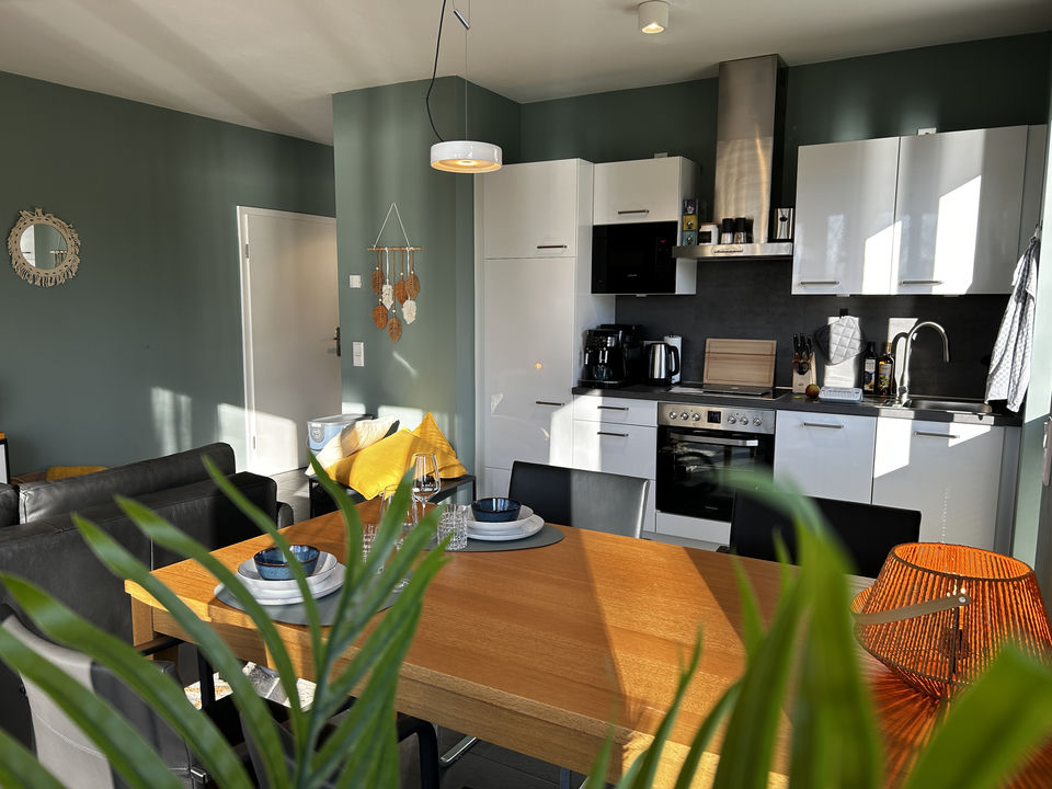 A wooden dining table set with blue dishes stands in a sunlit open-plan kitchen with white cabinets and sage green walls.