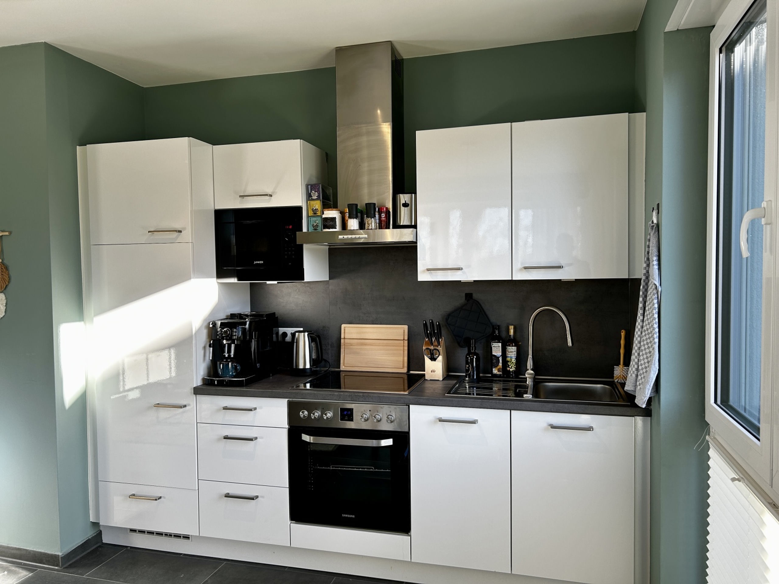 Compact white kitchen with glossy cabinets, dark countertop, built-in oven, induction hob, coffee machine, kettle, and a stainless extractor hood against a green wall.