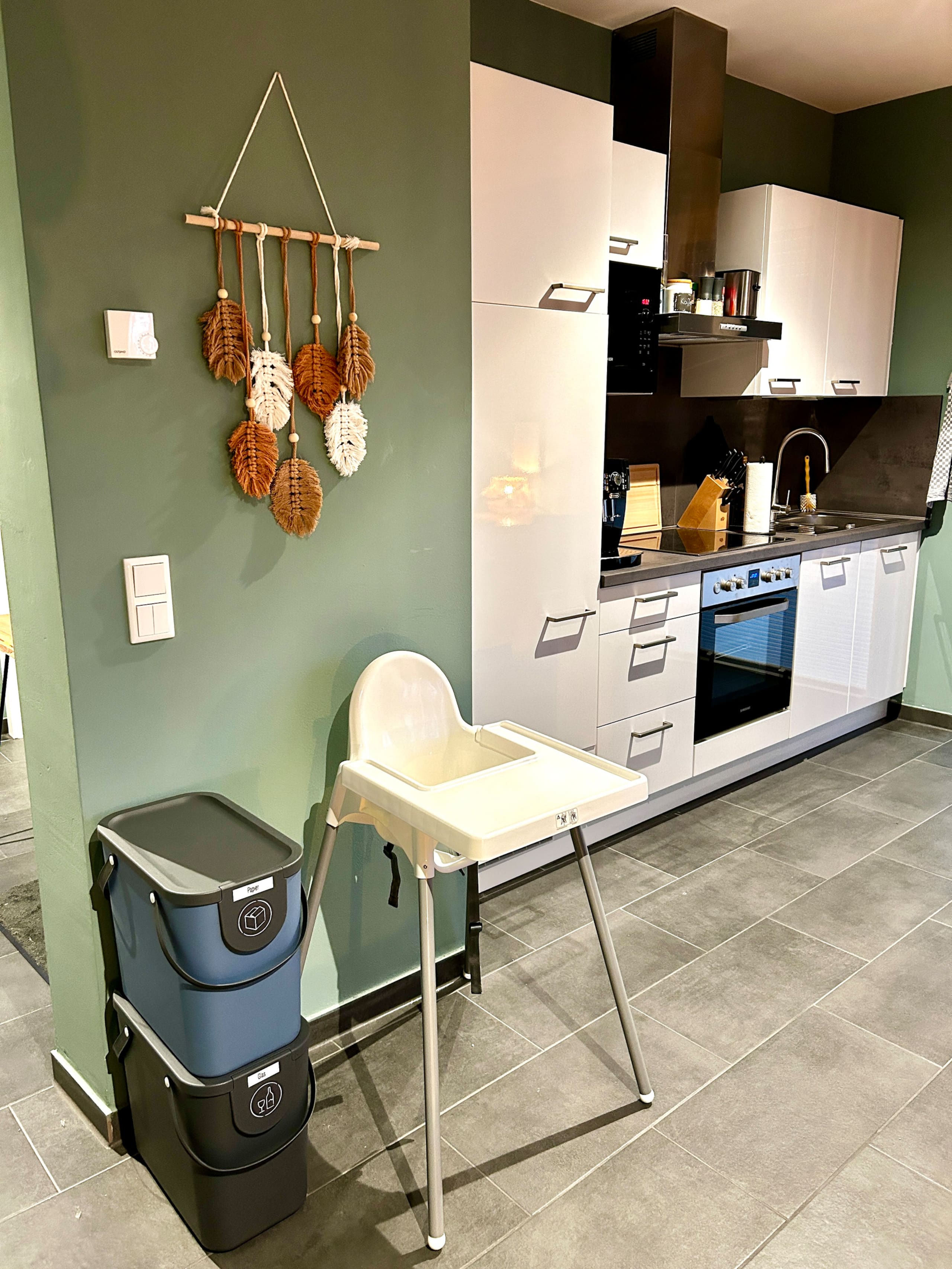 A white high chair with tray stands next to stacked recycling bins under a macramé leaf hanging; the kitchen glows warmly in the background.