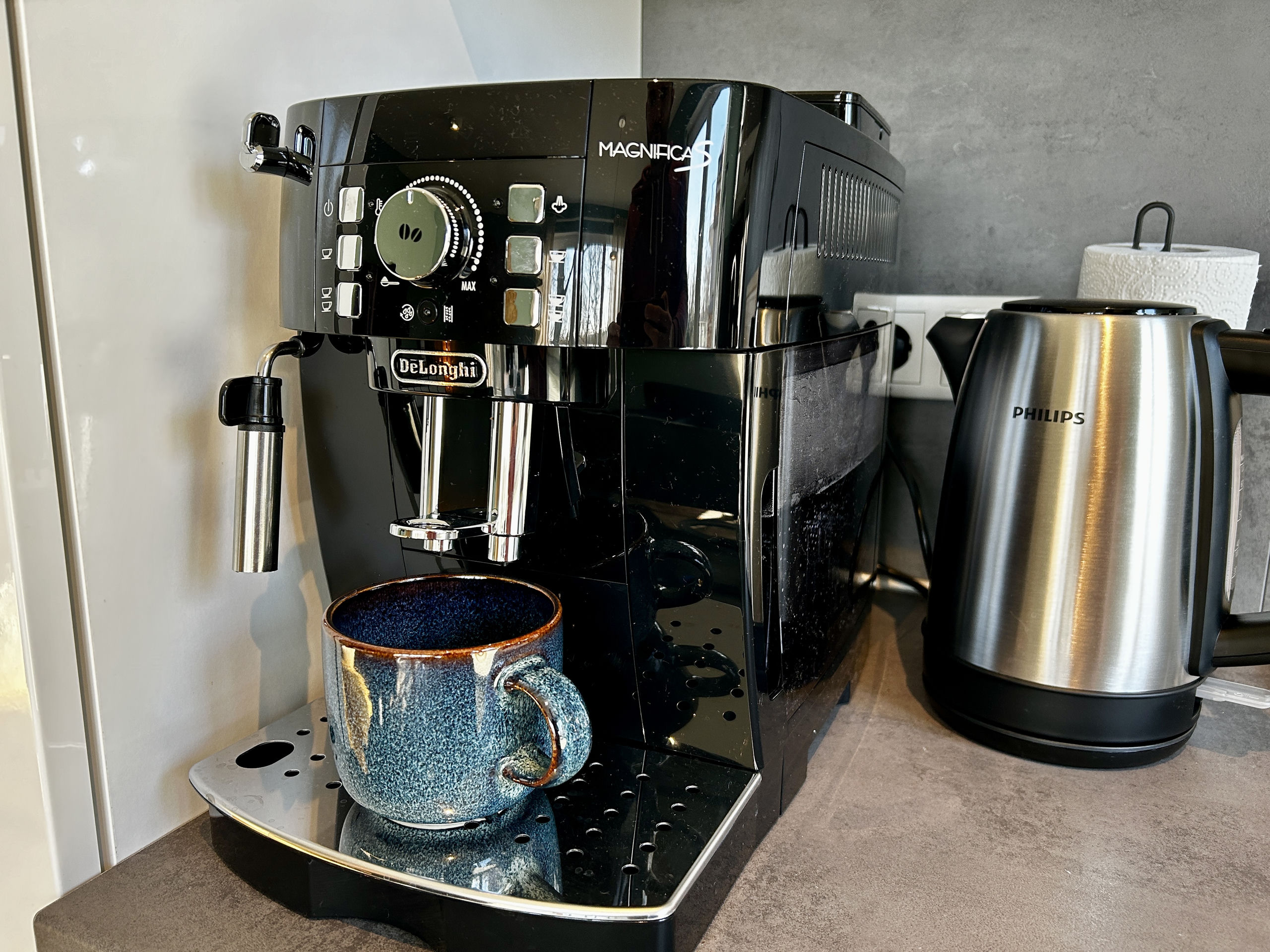 Close-up of a black bean-to-cup coffee machine with a blue glazed mug on the drip tray and a steel kettle beside it.