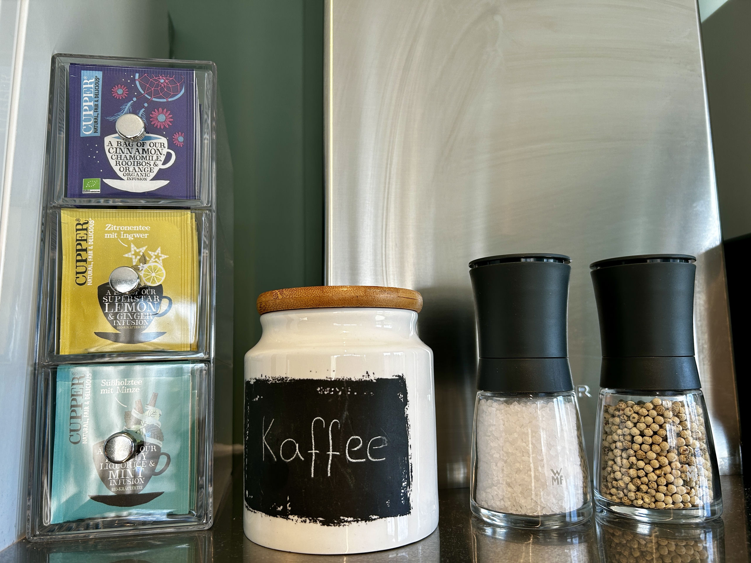 Tiered acrylic tea organizer with Cupper tea packets, a chalkboard-labelled coffee jar, and two electric grinders sit beside a steel extractor hood.