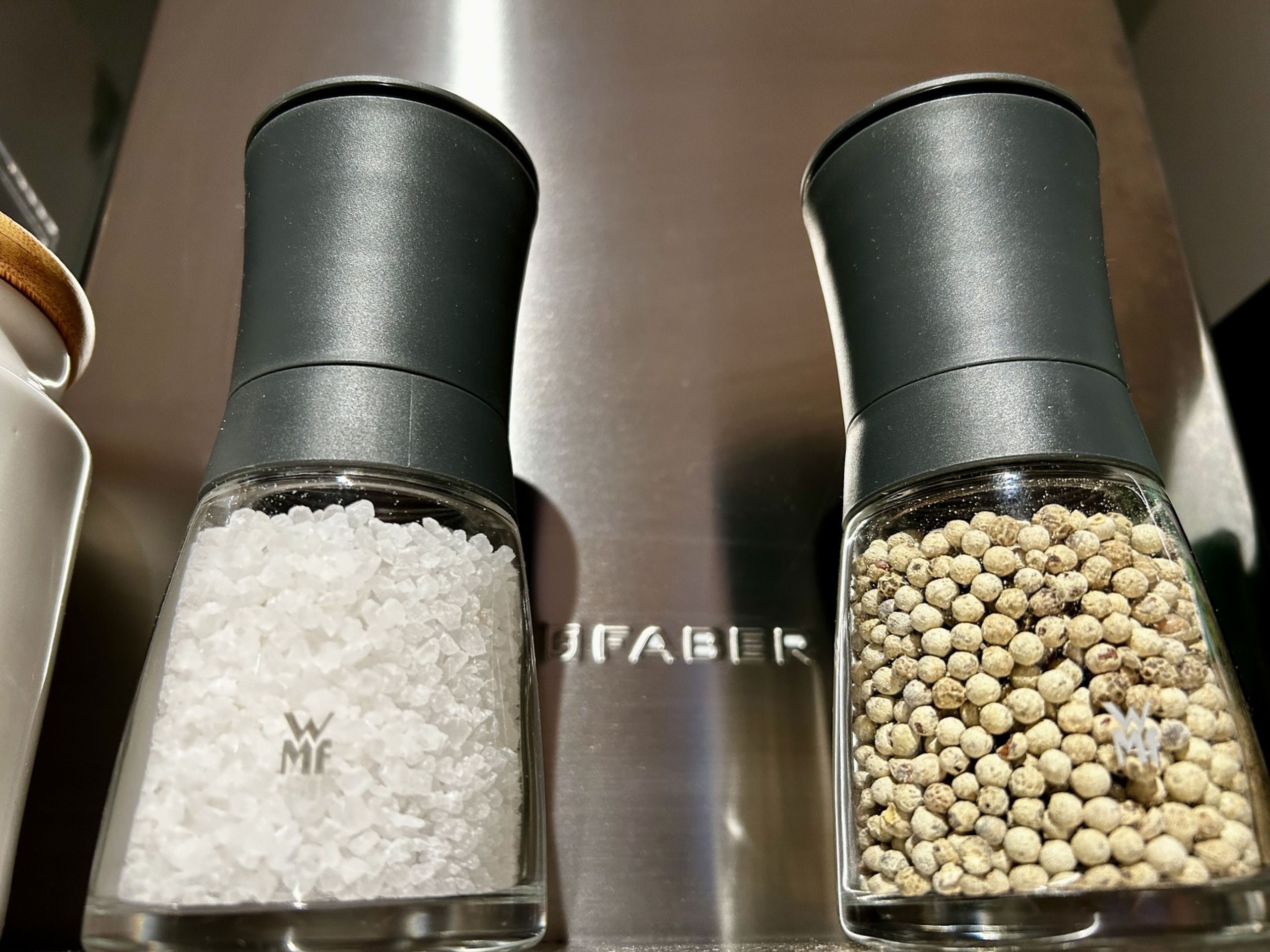Macro shot of two electric WMF grinders filled with coarse salt and white peppercorns in front of a brushed steel surface.