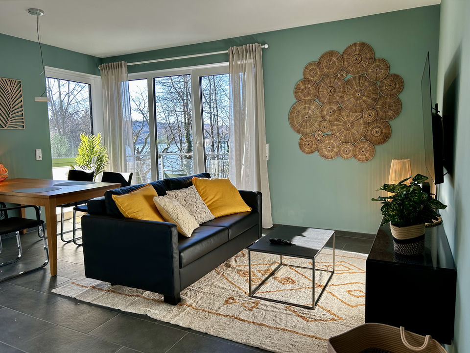 Sunlit living room with a black leather sofa, yellow cushions, rattan wall art, and a lake glimpse through the balcony doors.