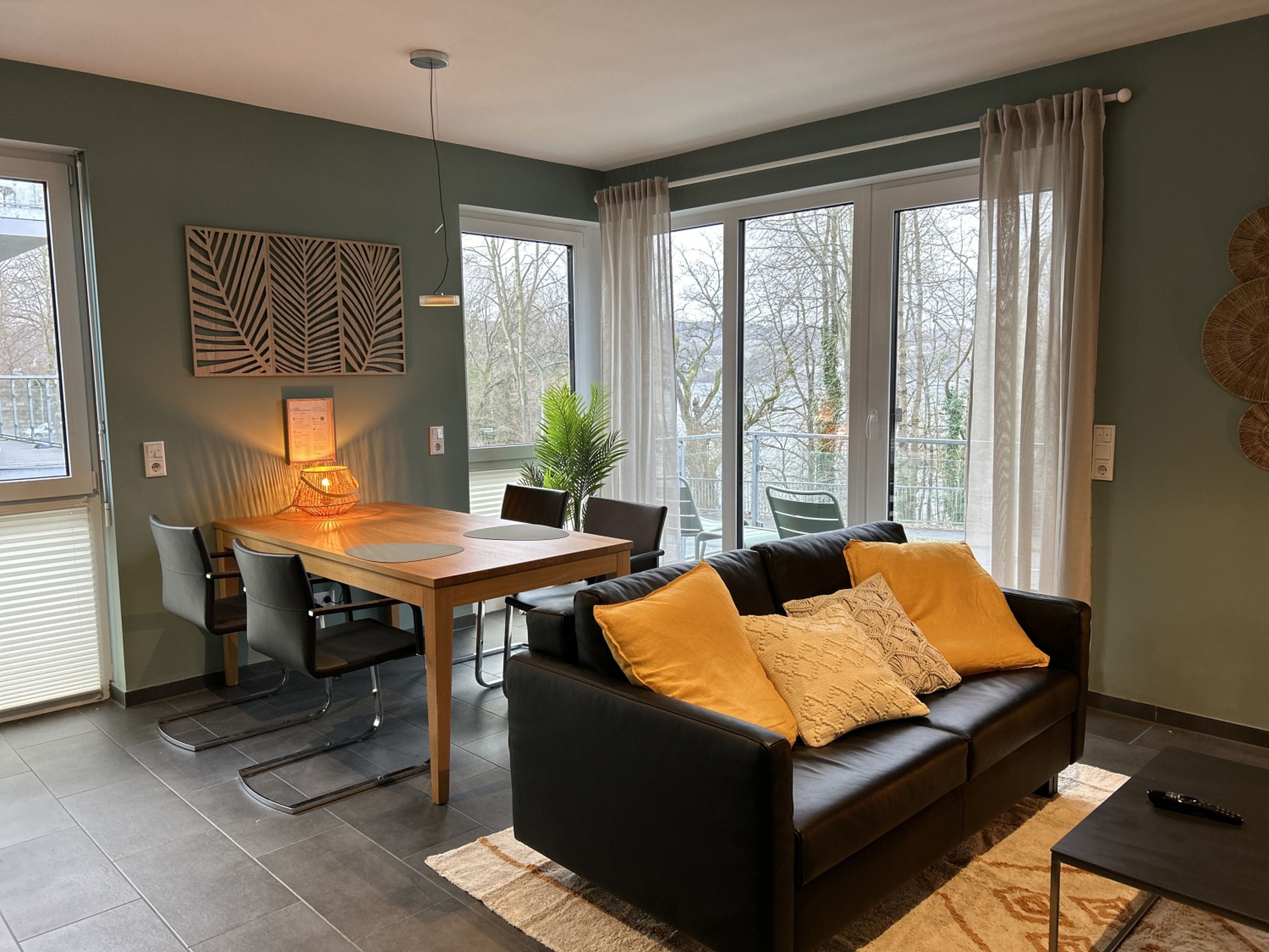 Evening scene of the dining area with a wooden table, four black chairs, warm rattan lamp, and balcony doors at dusk.