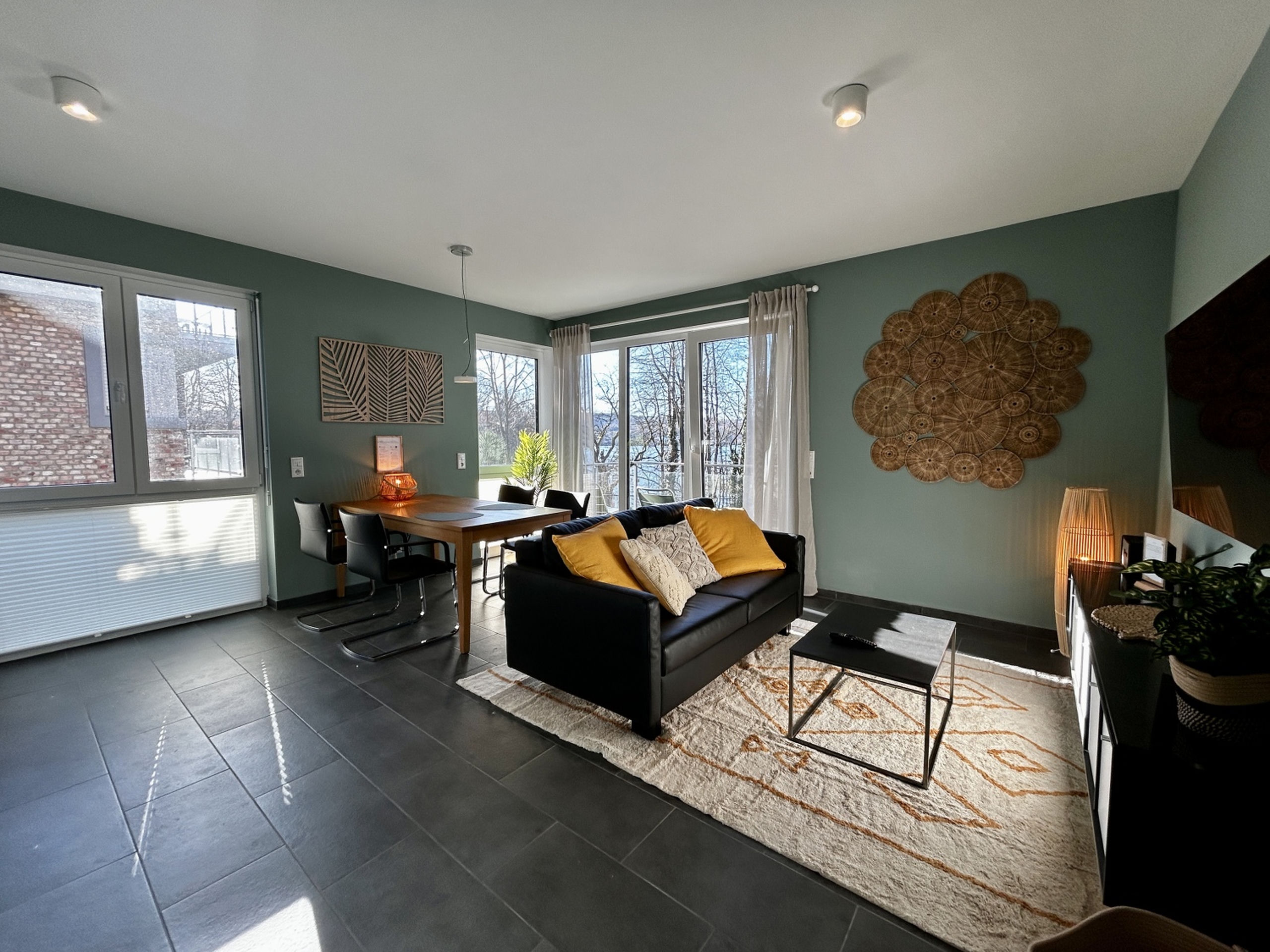 Wide view of the open-plan living and dining room with grey tiled floor, sage walls, rattan wall art, and balcony doors.