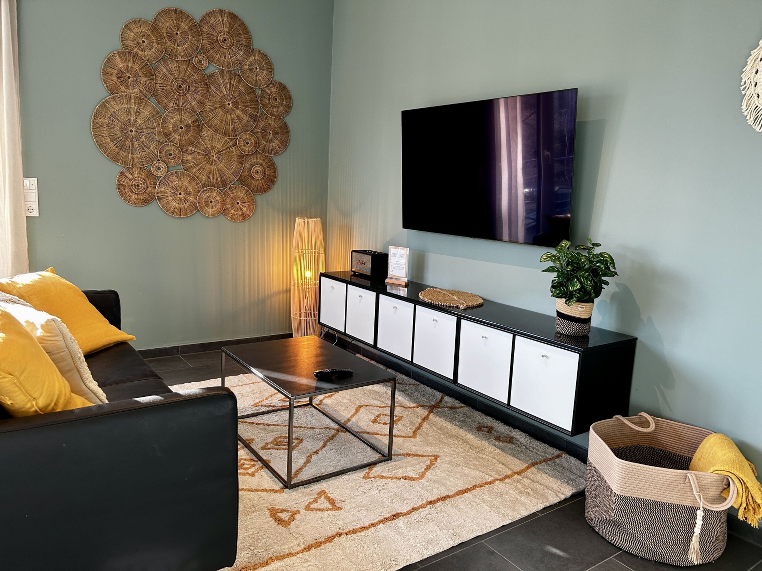 Media wall of the sage living room seen from the sofa side, with rattan sunburst wall art, a wall-mounted TV above a black sideboard with white cube inserts, a candle-like rattan floor lamp, and the corner of a black leather sofa with yellow cushions.