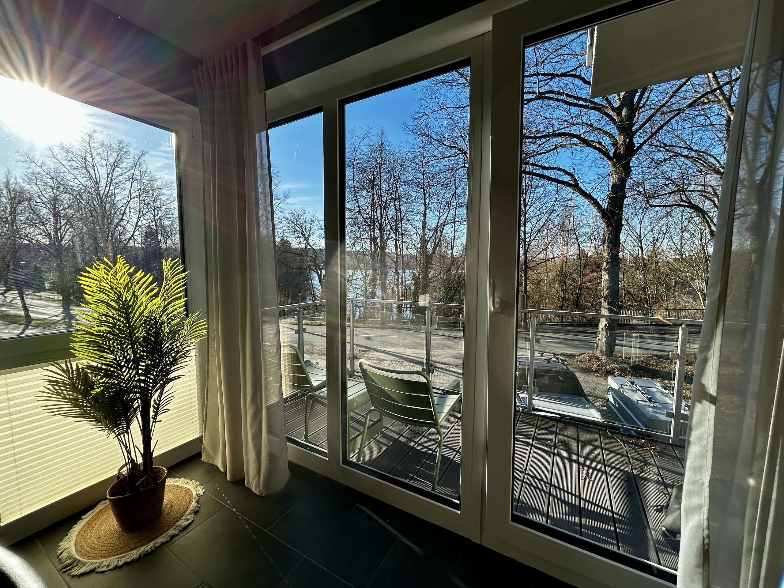 Backlit balcony corner where sunlight streams through tall windows, highlighting a potted palm and the lake view beyond the railing.