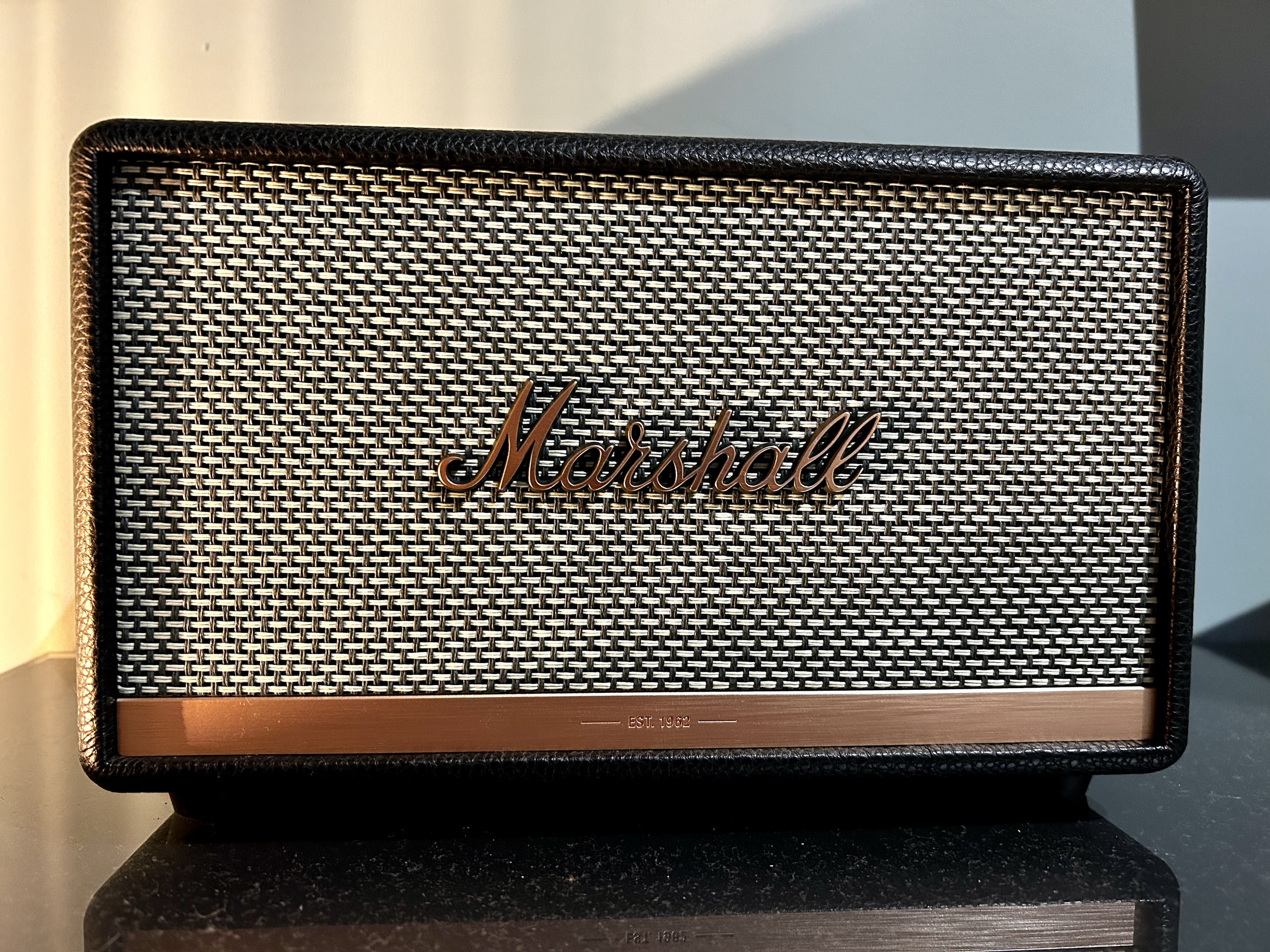 Close-up of a Marshall Bluetooth speaker with woven grille and copper lettering, standing on a dark shelf in warm light.