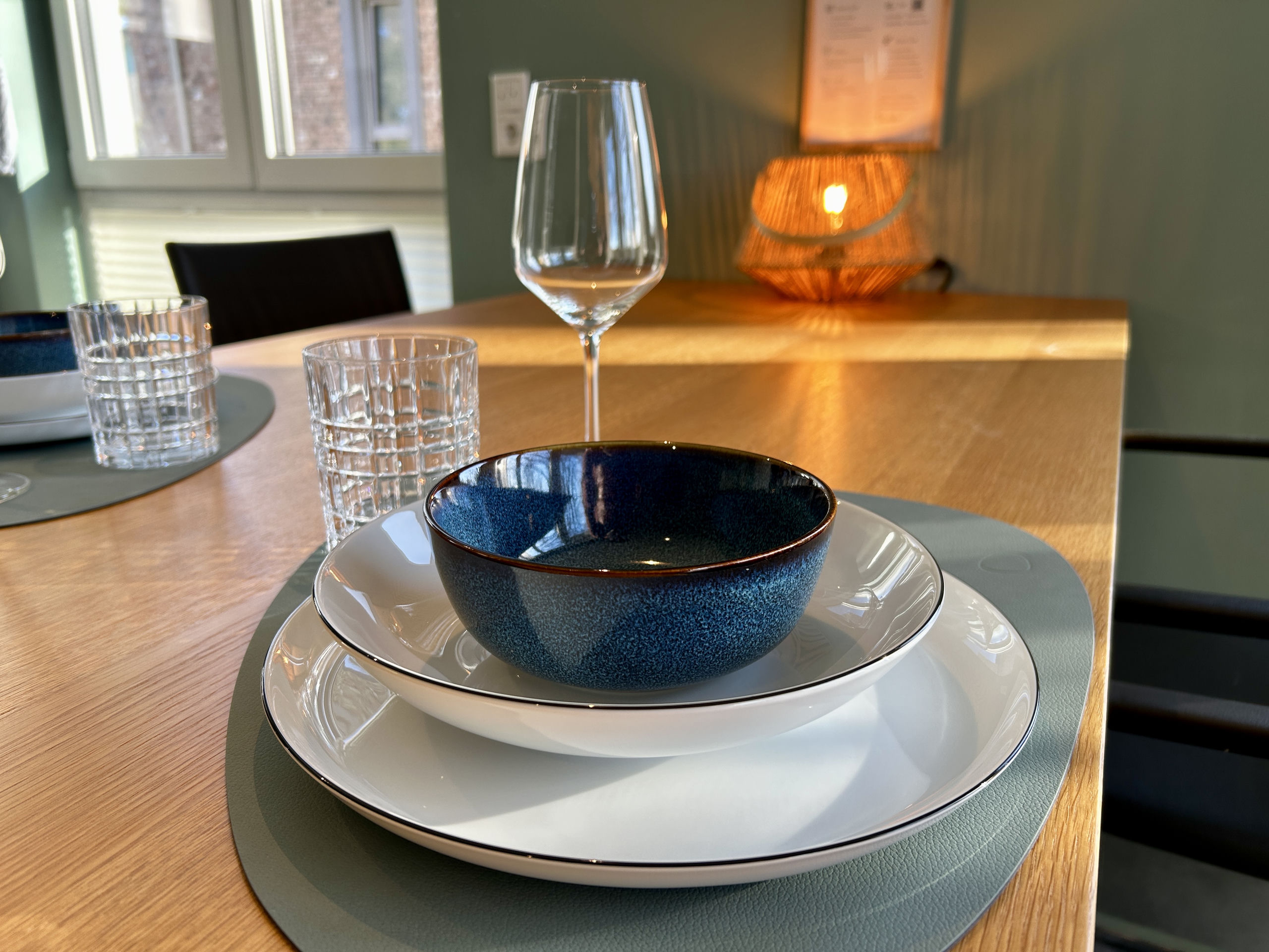 Table setting on a wooden dining table with a blue-glazed bowl, white plates, crystal glasses, and a glowing rattan lamp behind.