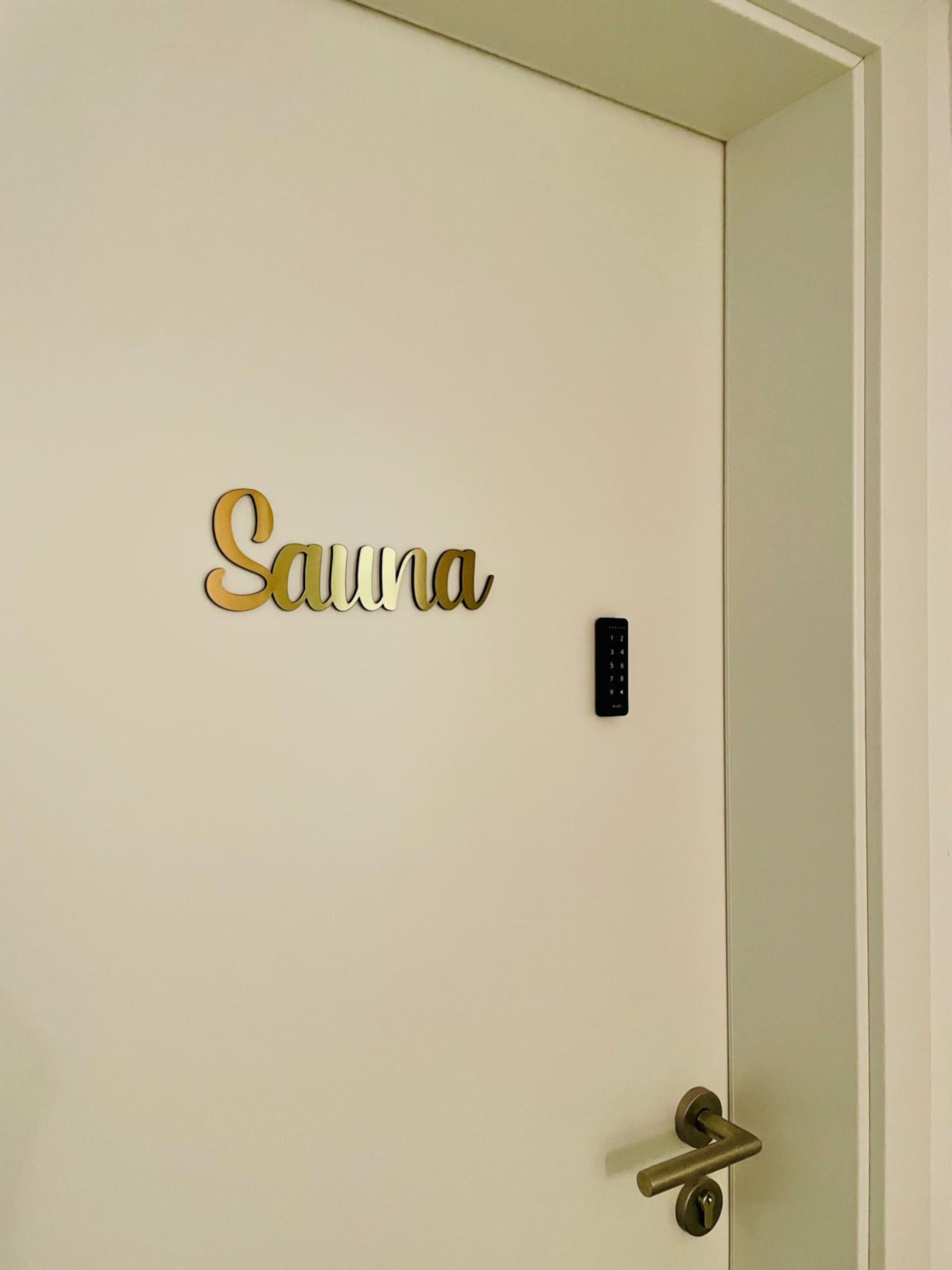 White interior door with a gold cursive Sauna sign, a small black numeric keypad, and a brass handle.