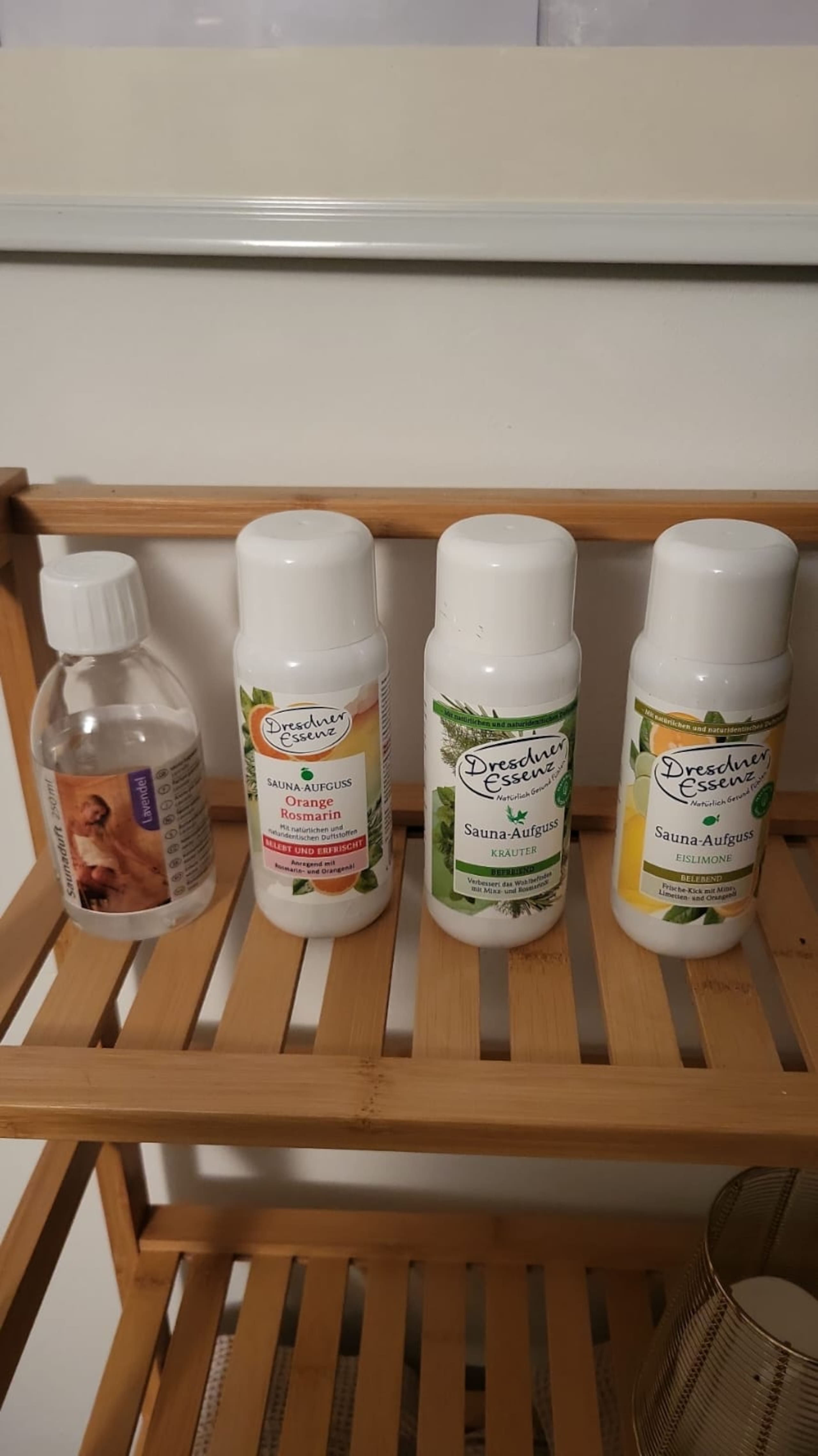 Four sauna additives on a bamboo shelf: a small lavender scent bottle and three Dresdner Essenz Aufguss concentrates in orange-rosemary, herbs, and iced lemon.