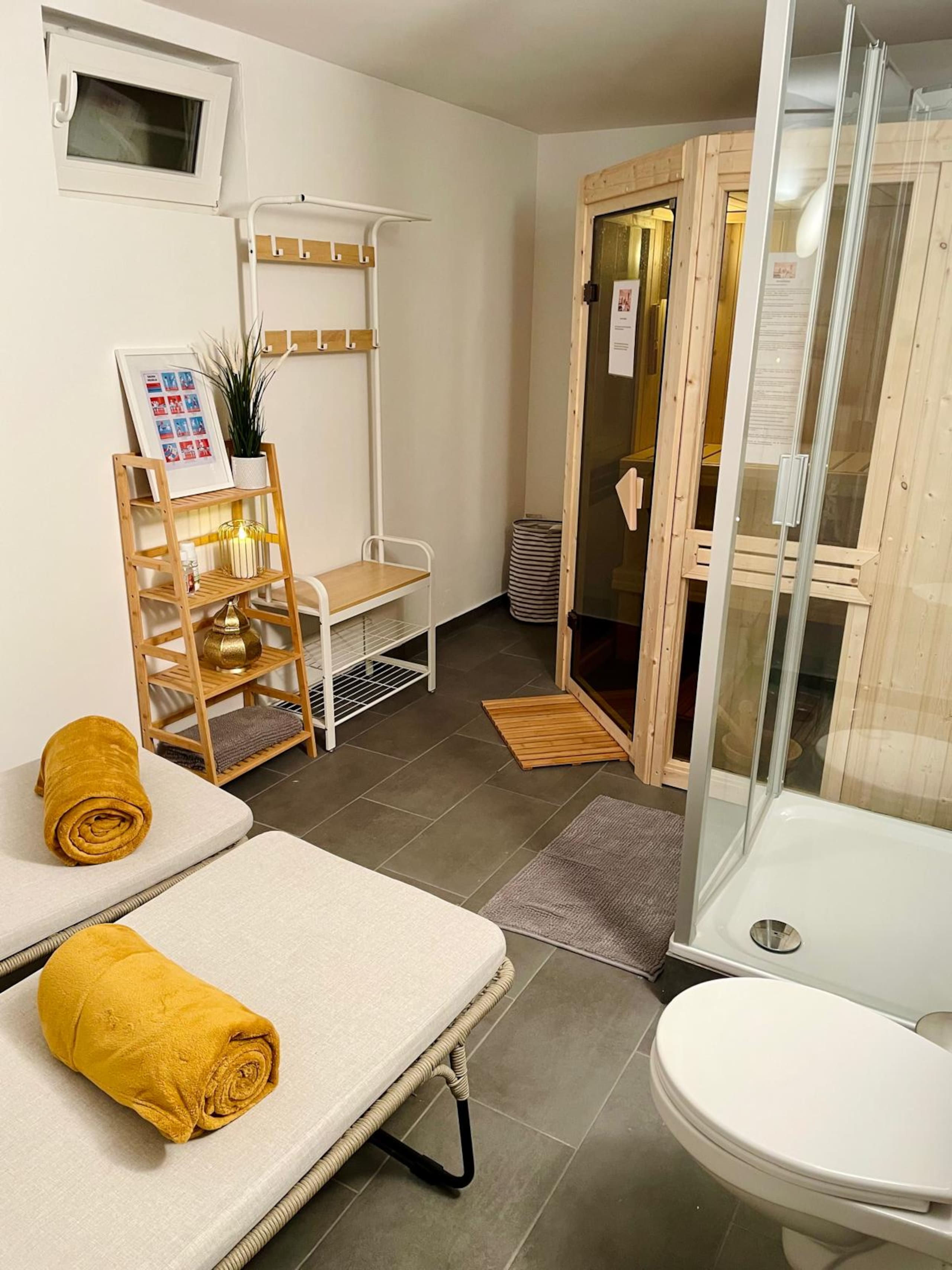 Spa corner with two loungers holding rolled mustard towels, an open wooden sauna cabin, a glass shower, and a toilet.