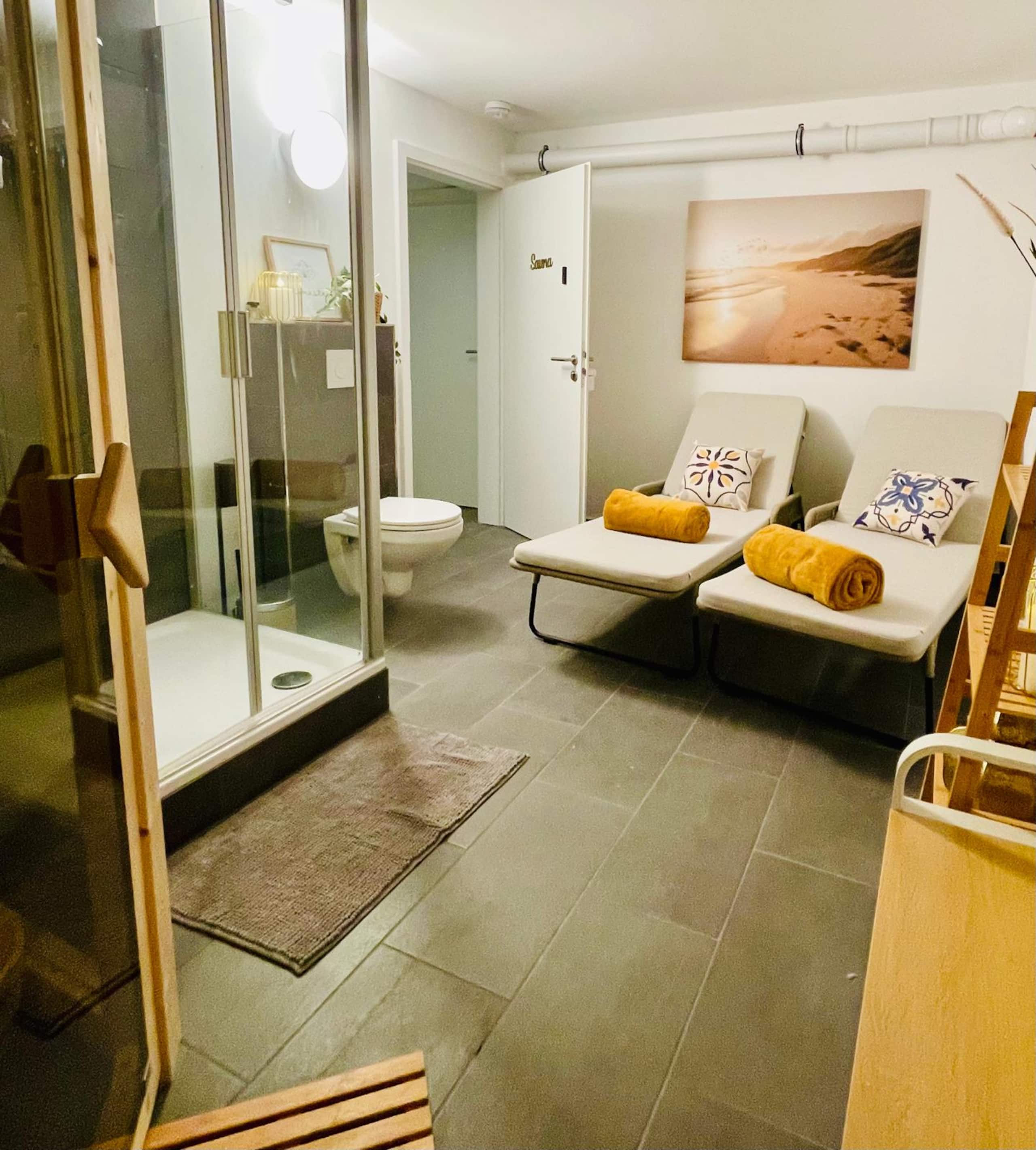 Relax area with two loungers and floral cushions below a beach photograph, the shower and the sauna door behind them.