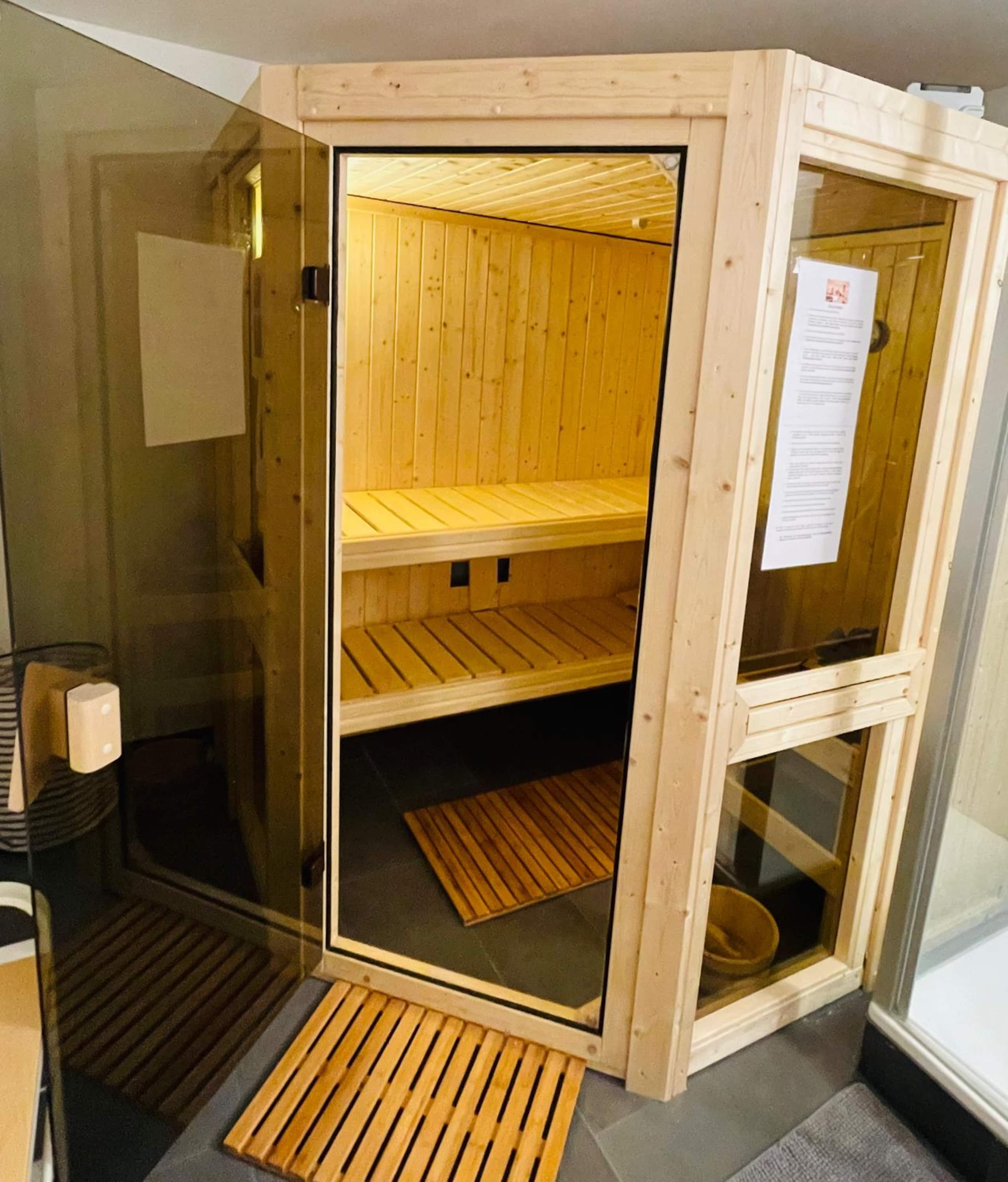 Compact corner sauna cabin in light wood with its glass door open, showing two empty benches and wooden floor mats.