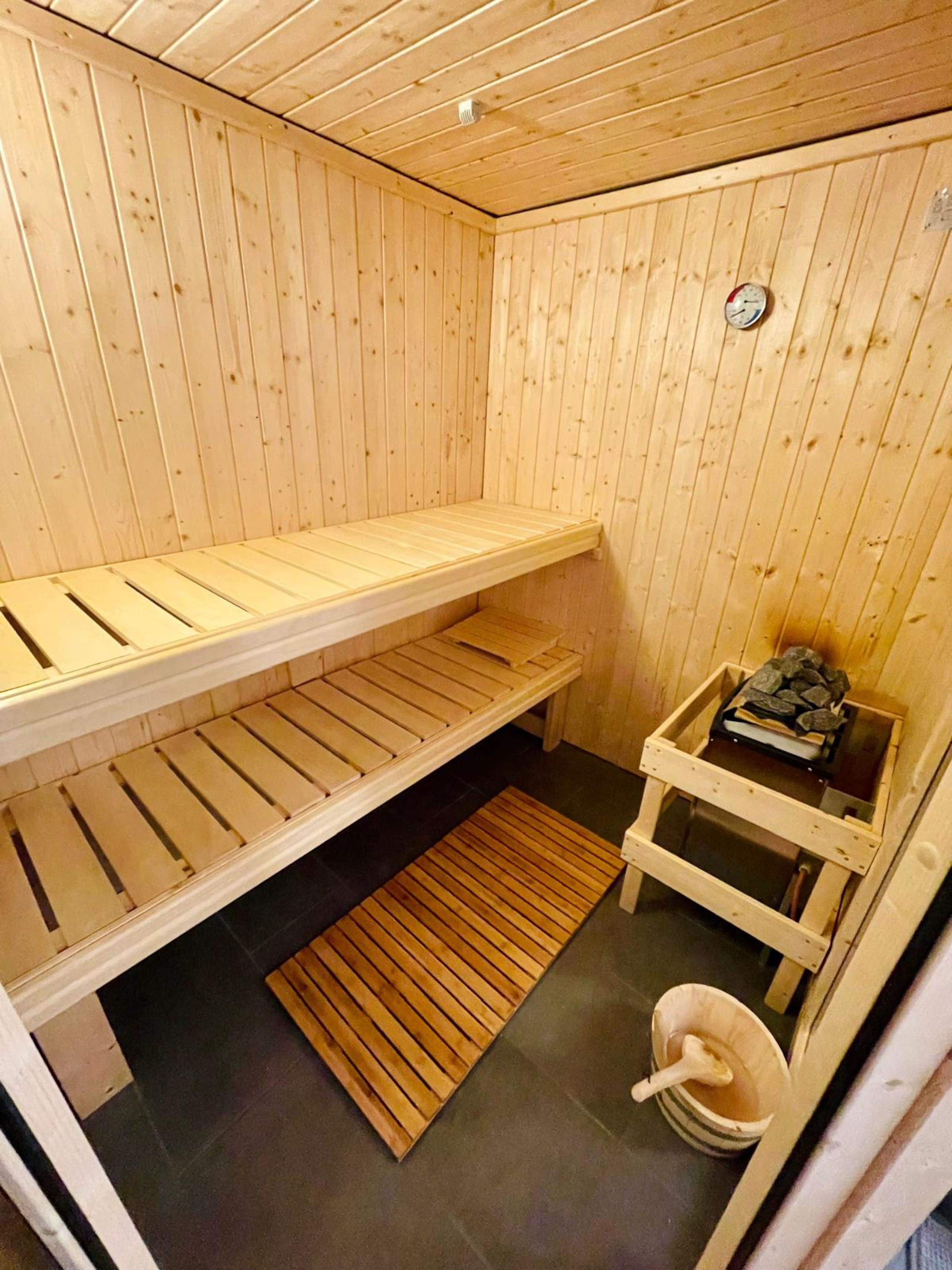 Inside the sauna: two wooden benches, a stone-filled heater, a round wall thermometer, and a wooden bucket with a ladle.