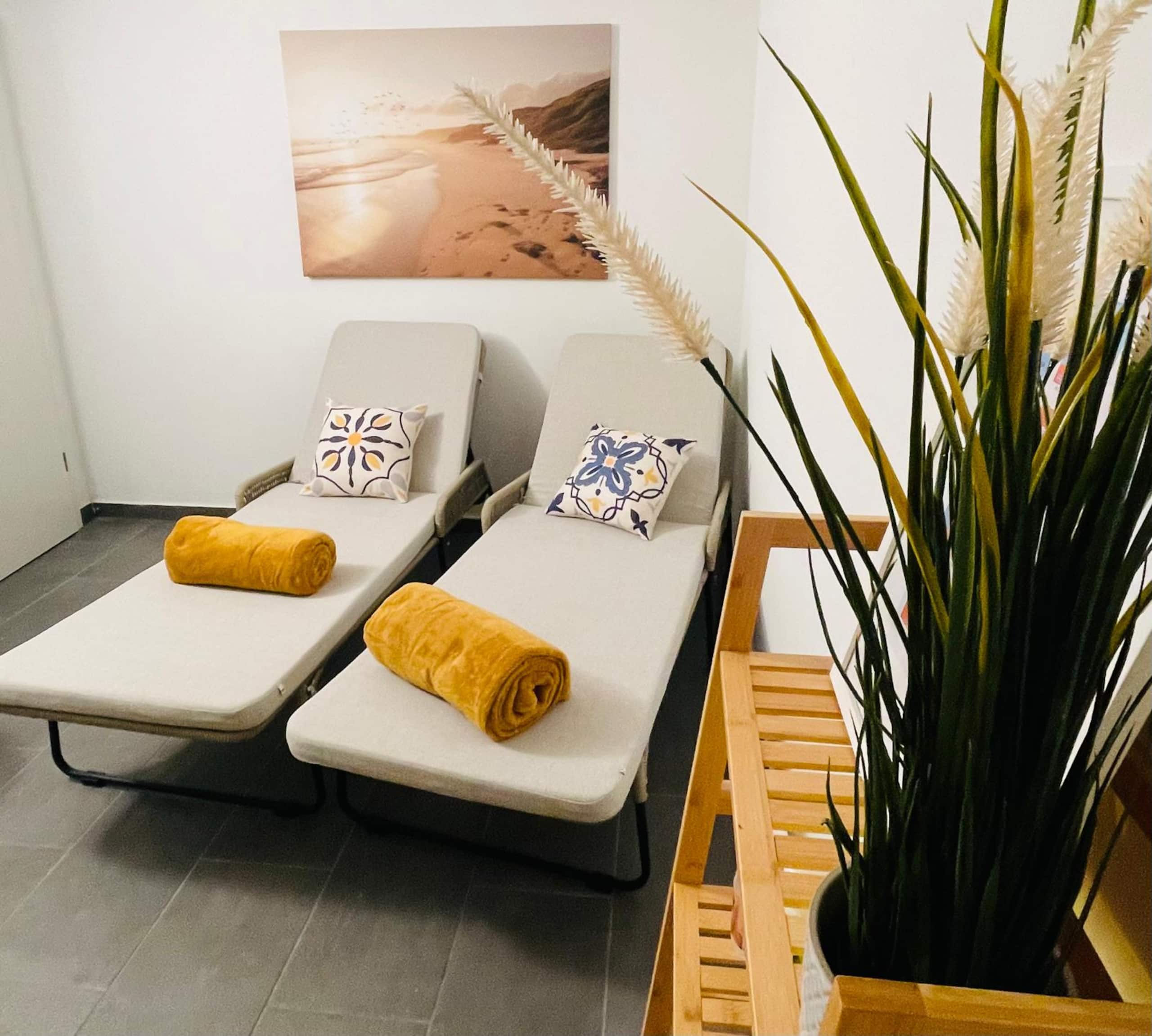 Two relax loungers with patterned cushions and rolled mustard towels, a beach photograph above and pampas grass in the foreground.