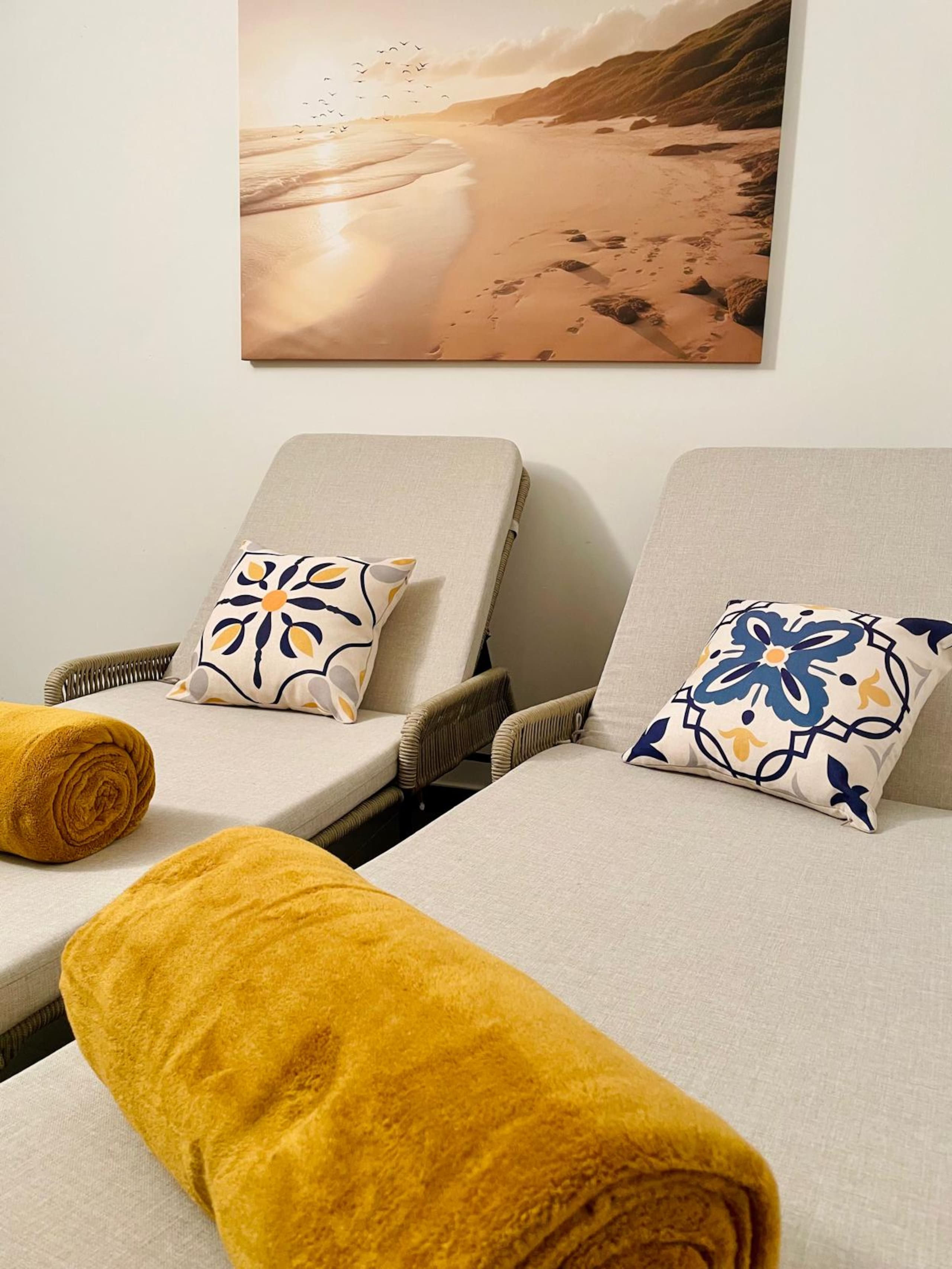 Close view of the two relax loungers with their floral cushions and rolled towels beneath the sandy-bay wall canvas.