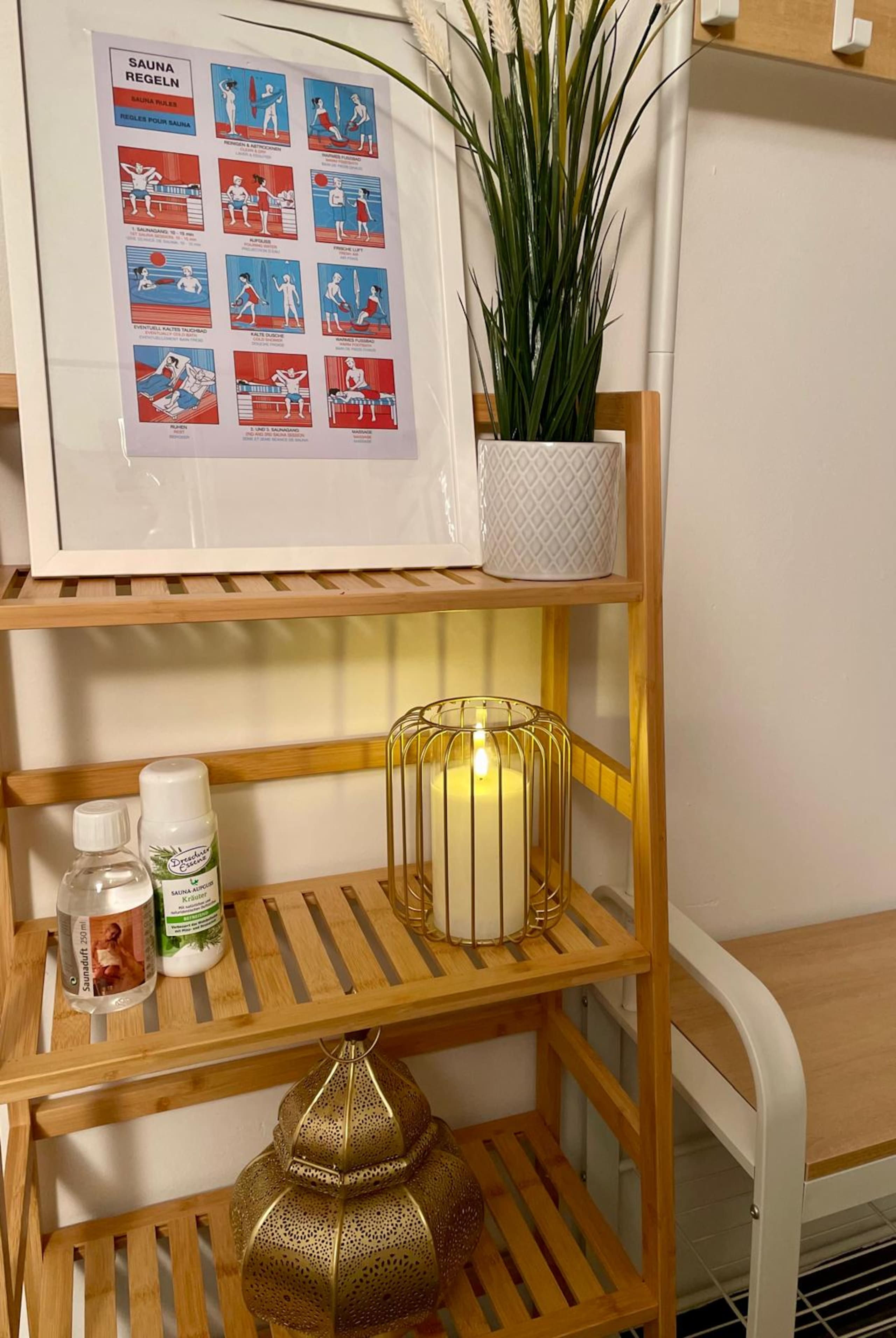 Bamboo shelf with a framed illustrated sauna-rules poster, a plant, a lit candle in a gold cage, and sauna infusion bottles.