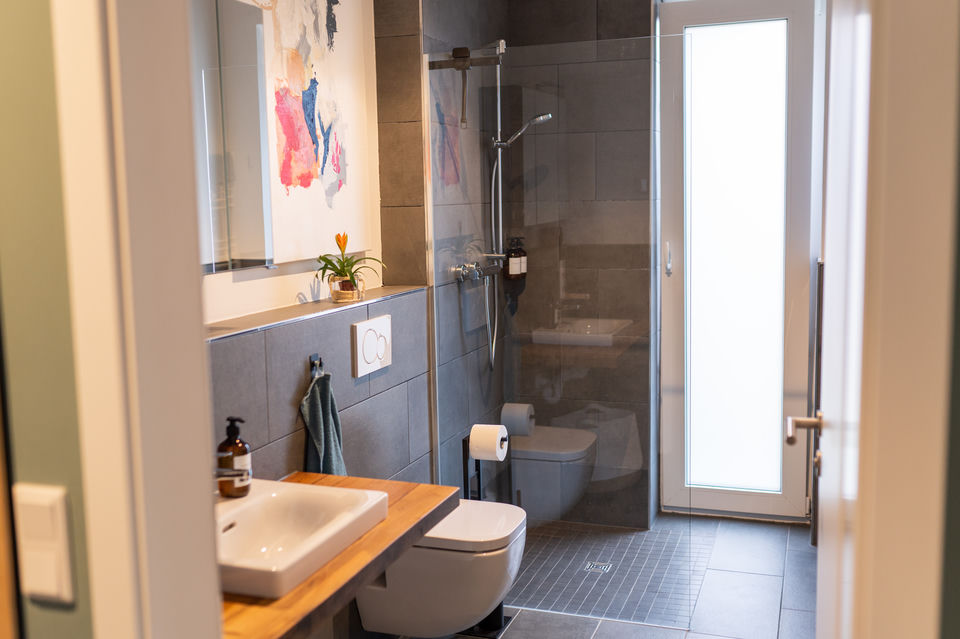 Bagno moderno con piastrelle grigie alle pareti, doccia a filo con parete in vetro, WC sospeso e mobile in legno con lavabo bianco rettangolare, con un dipinto astratto colorato riflesso nello specchio.