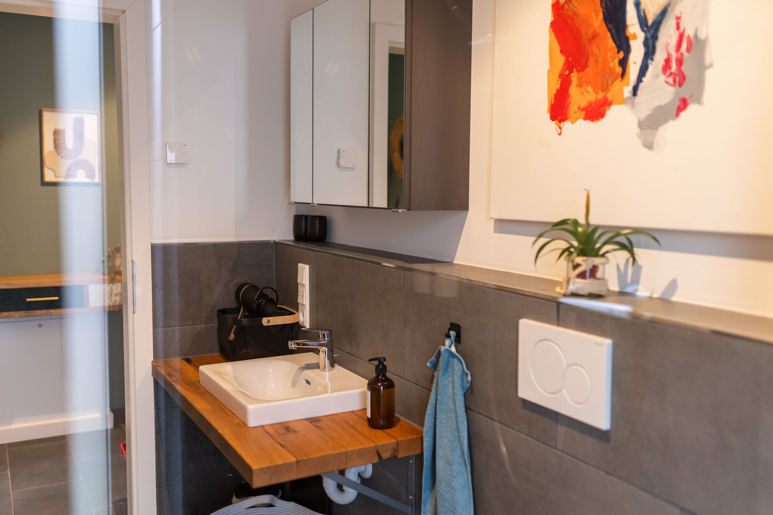 Zweites Waschplatz-Ensemble mit schwebendem Holzwaschtisch, weißem Becken, Spiegelschrank und großformatigen grauen Fliesen; ein orangefarbenes abstraktes Bild hängt an der Wand, die Tür führt ins Schlafzimmer.