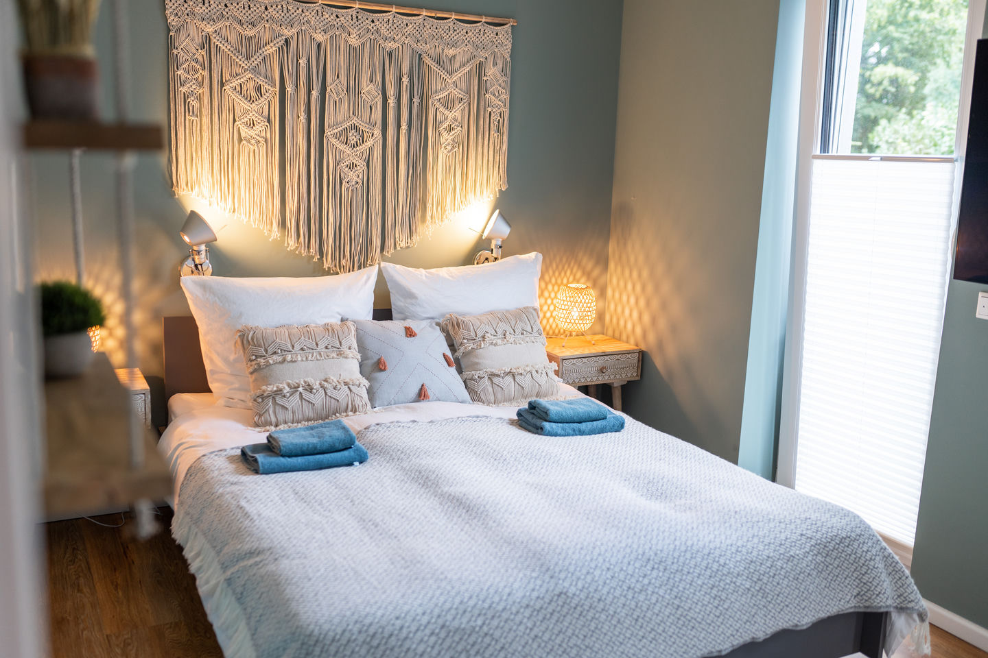 Cama de matrimonio con ropa de cama blanca, colcha tejida gris y cojines decorativos crema, iluminada por dos lámparas de lectura bajo un gran tapiz de macramé en una pared verde.