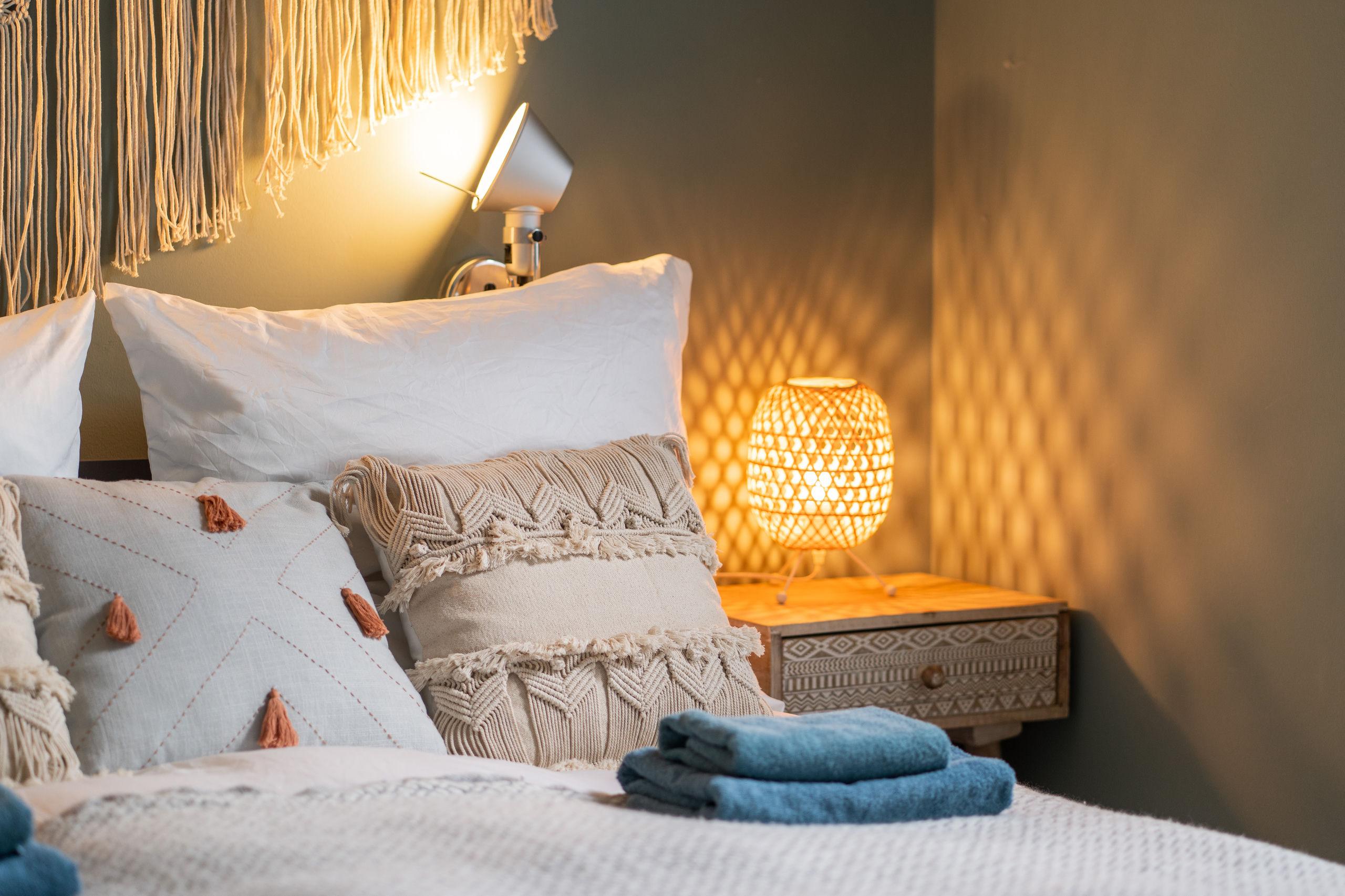 Nahaufnahme des gedeckten Betts mit cremefarbenen Makramee- und Quasten-Kissen, einer silbernen Klemm-Leselampe und einer Rattan-Tischlampe, die ein Muster an die Wand wirft.
