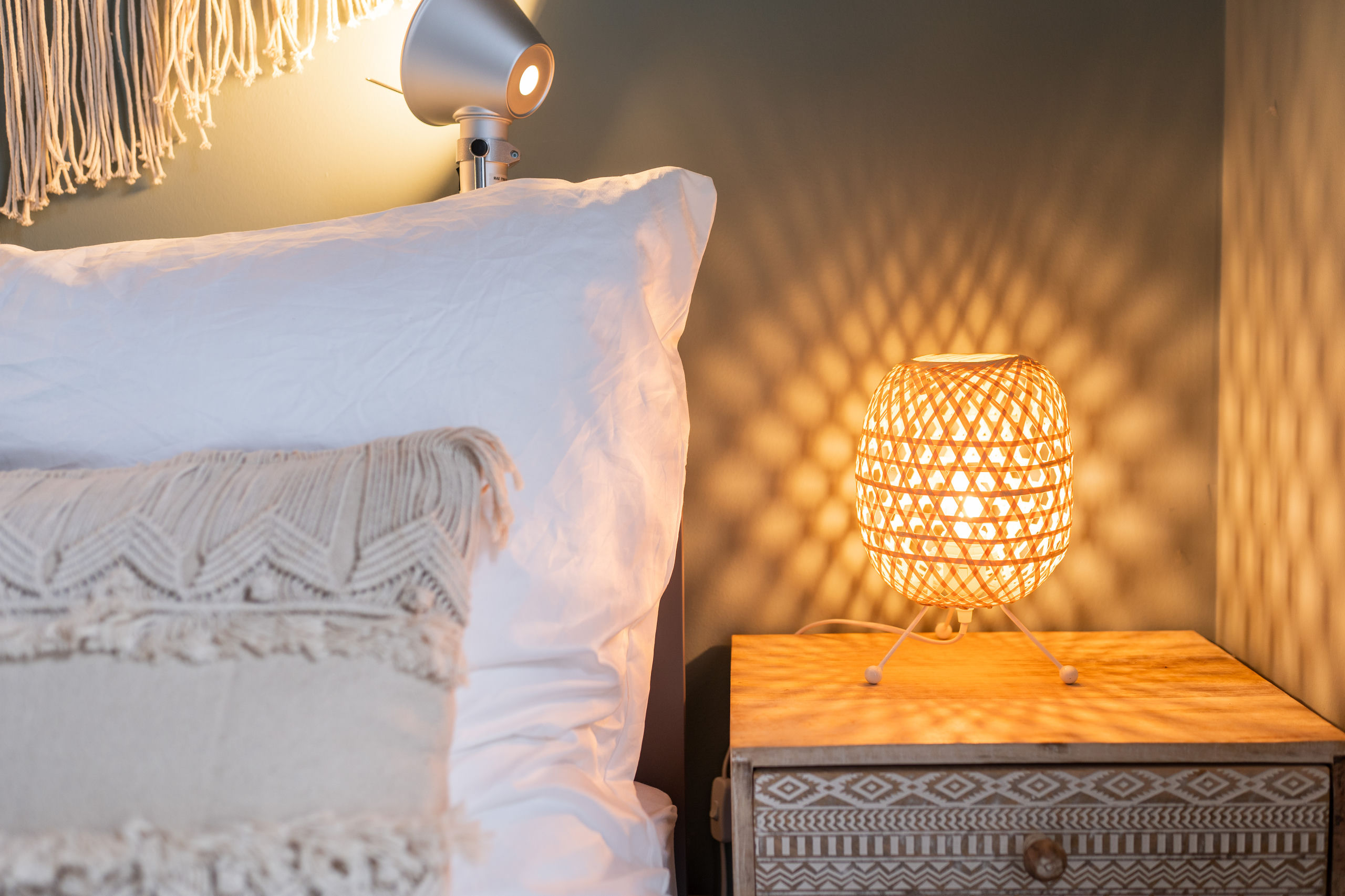 Detail einer leuchtenden Bambus-Nachttischlampe auf einem gemusterten Holz-Nachttisch neben weißen Kissen und einer silbernen Leselampe.