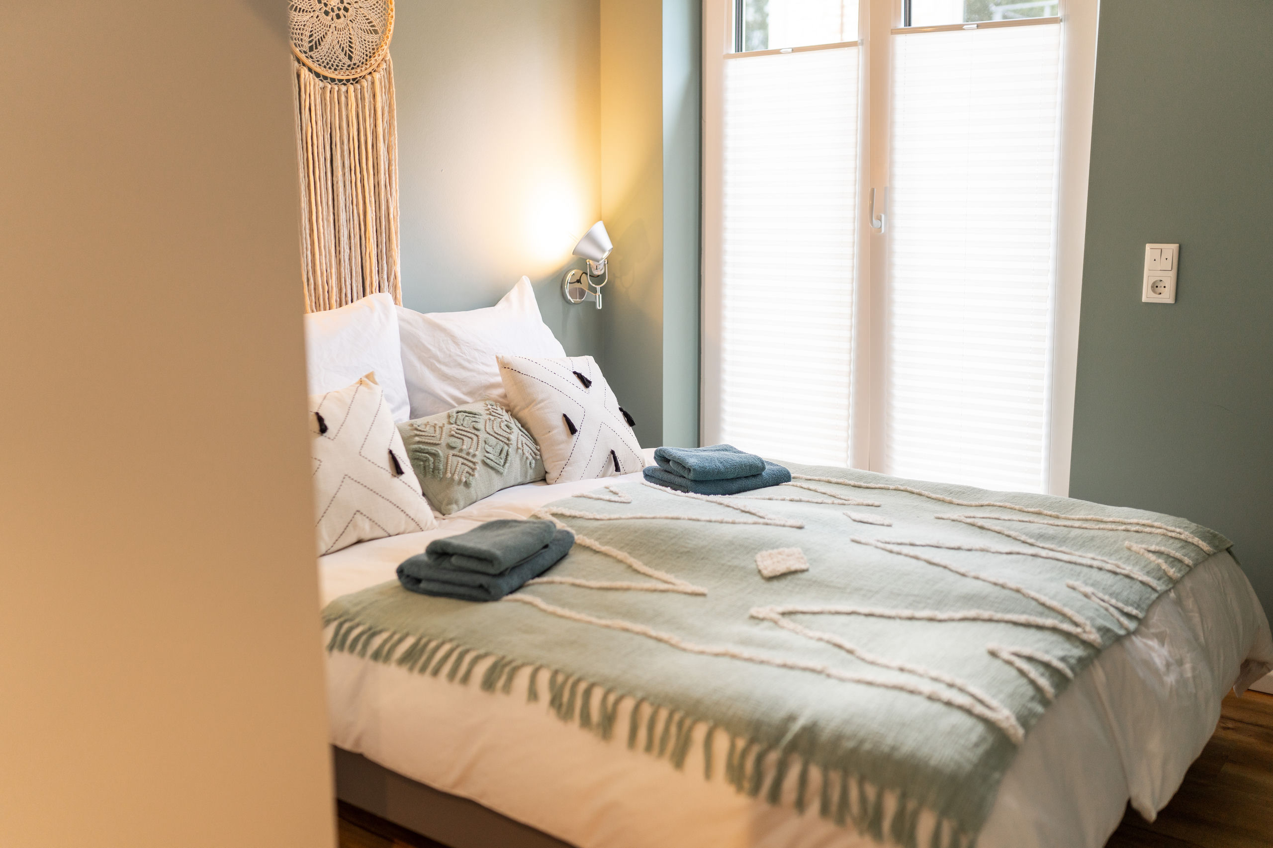 Zweites Schlafzimmer, durch die Tür gesehen, mit Doppelbett in salbeigrüner Chenille-Tagesdecke, weißen Kissen mit Quasten und rundem Makramee über dem Kopfteil.