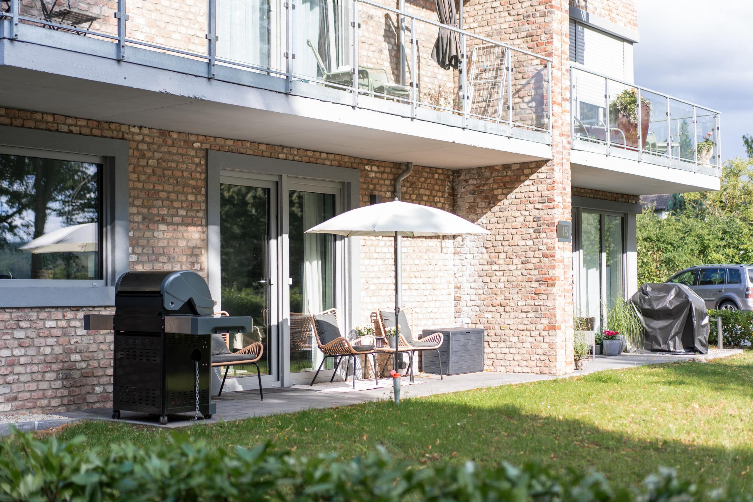Überdachte Terrasse des Backsteinhauses mit Rattanstühlen, weißem Sonnenschirm und Edelstahl-Gasgrill auf der gepflasterten Fläche vor den Terrassentüren.