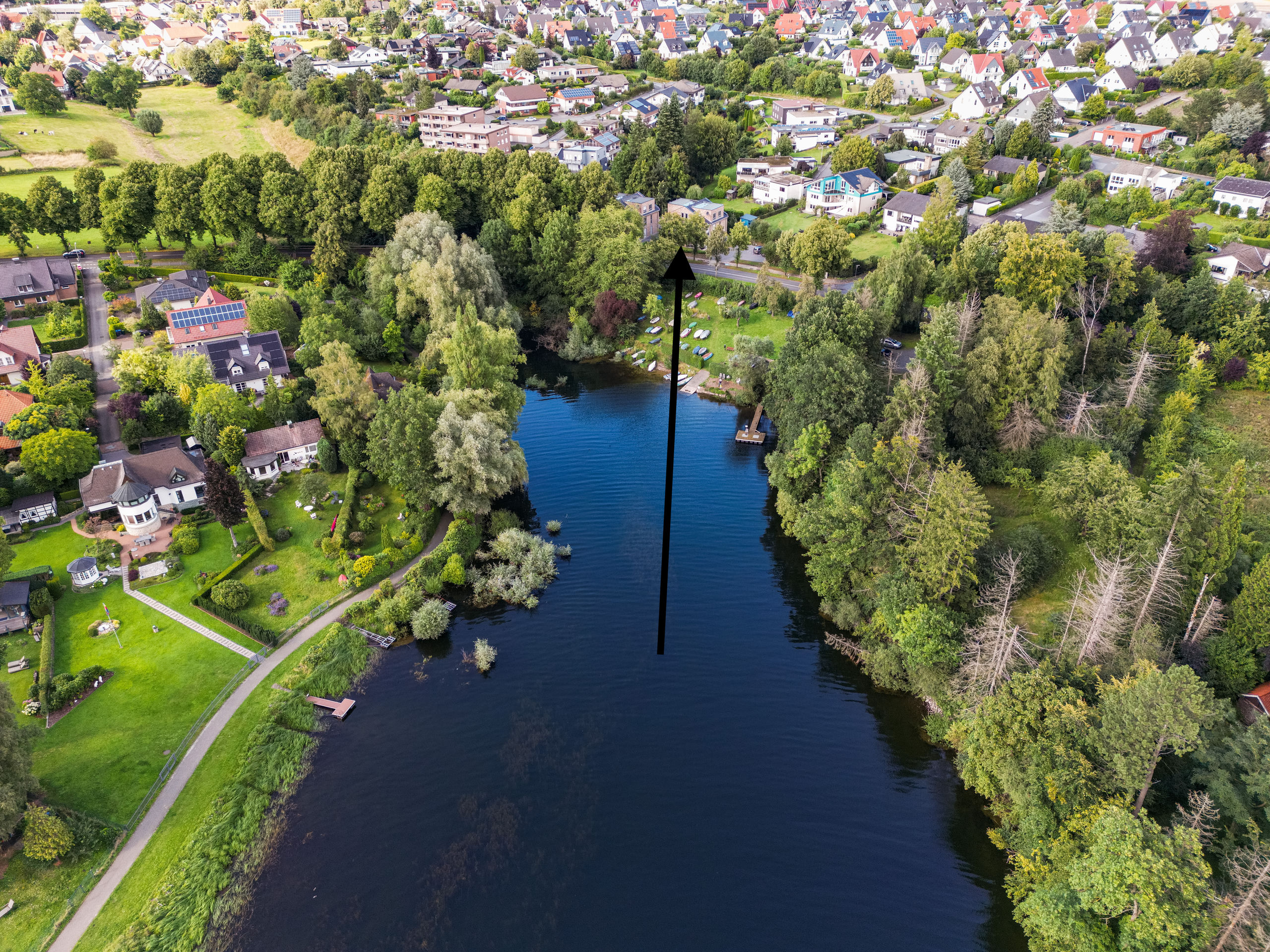 Luftaufnahme von der dunklen Wasserfläche aus in Richtung des baumgesäumten Ufers und der dahinterliegenden Ortschaft; ein langer schwarzer Pfeil zeigt auf das Land.