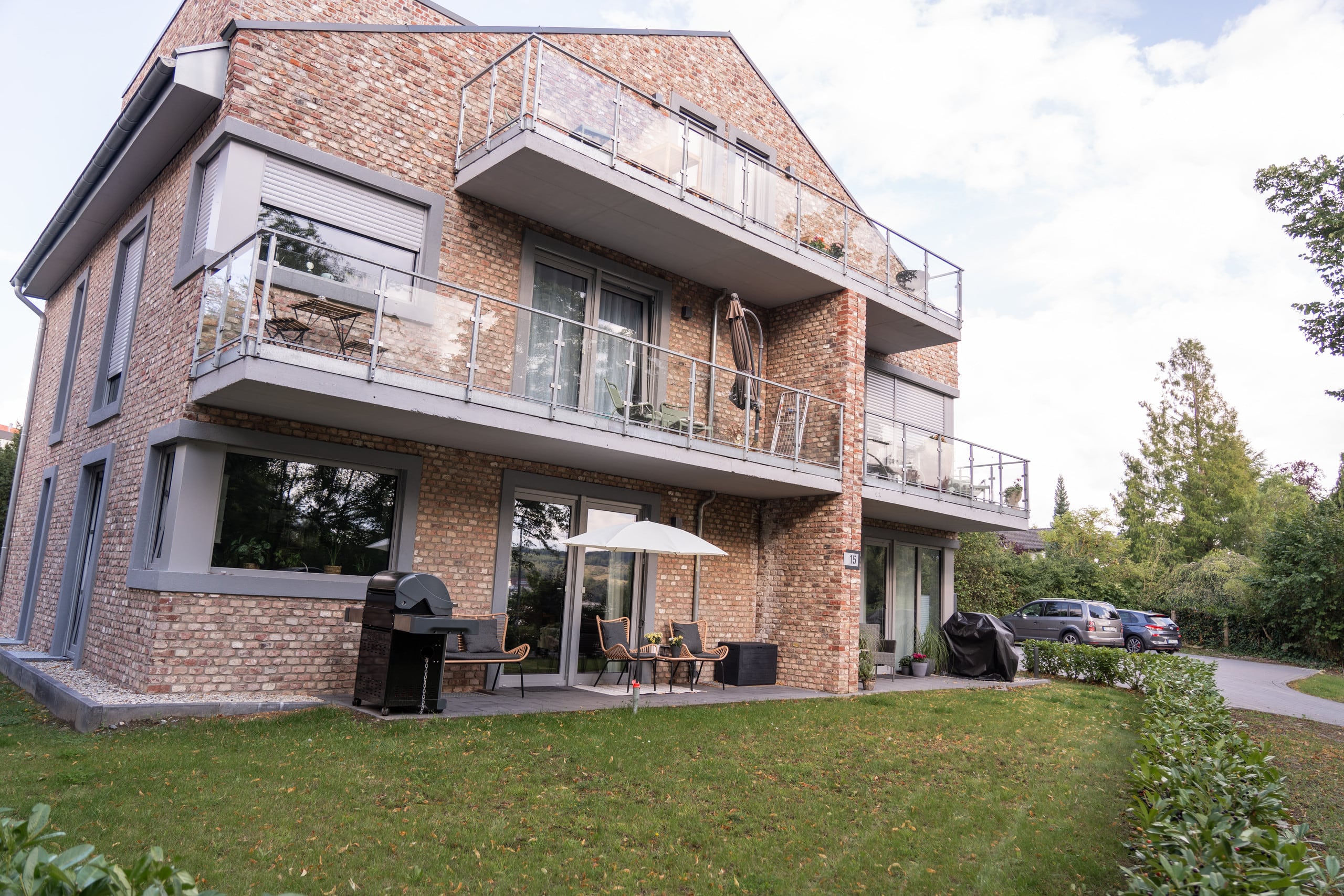 Dreigeschossiges Backsteinhaus vom Garten aus gesehen, mit Balkonen und Glasgeländern, Terrasse im Erdgeschoss und Rasen, der zur gemeinsamen Zufahrt führt.