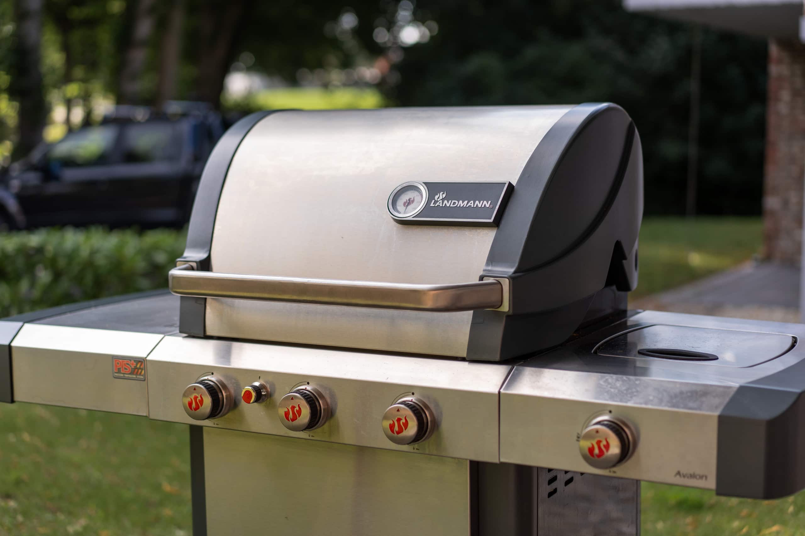 Nahaufnahme eines geschlossenen Edelstahl-Gasgrills mit vier Bediendrehknöpfen und Deckelthermometer, aufgestellt am Rand der Terrasse.