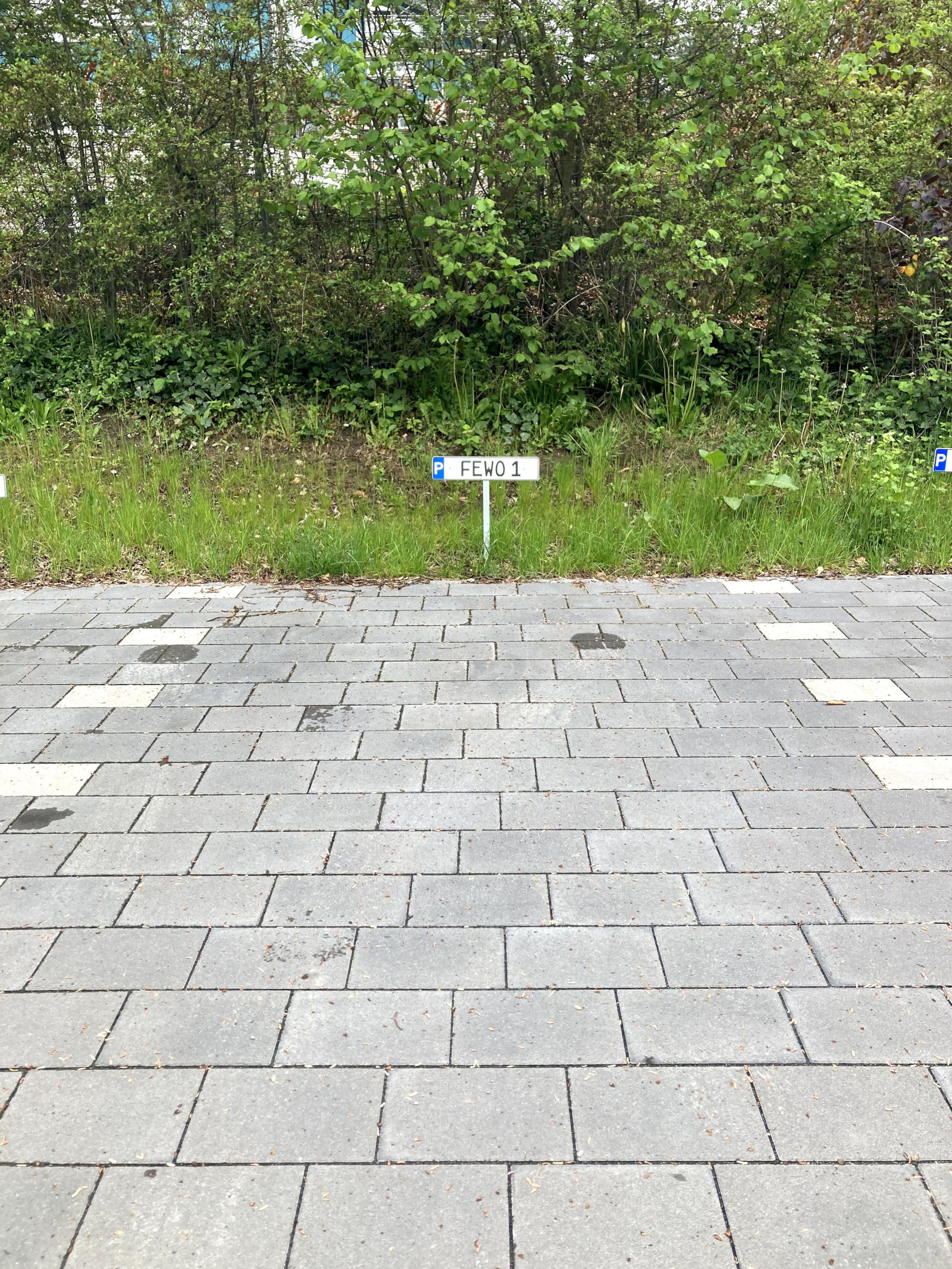 Gepflasterte Parkfläche mit kleinem Schild „P FEWO 1" am Rand einer geschnittenen Hecke, das den reservierten Stellplatz markiert.