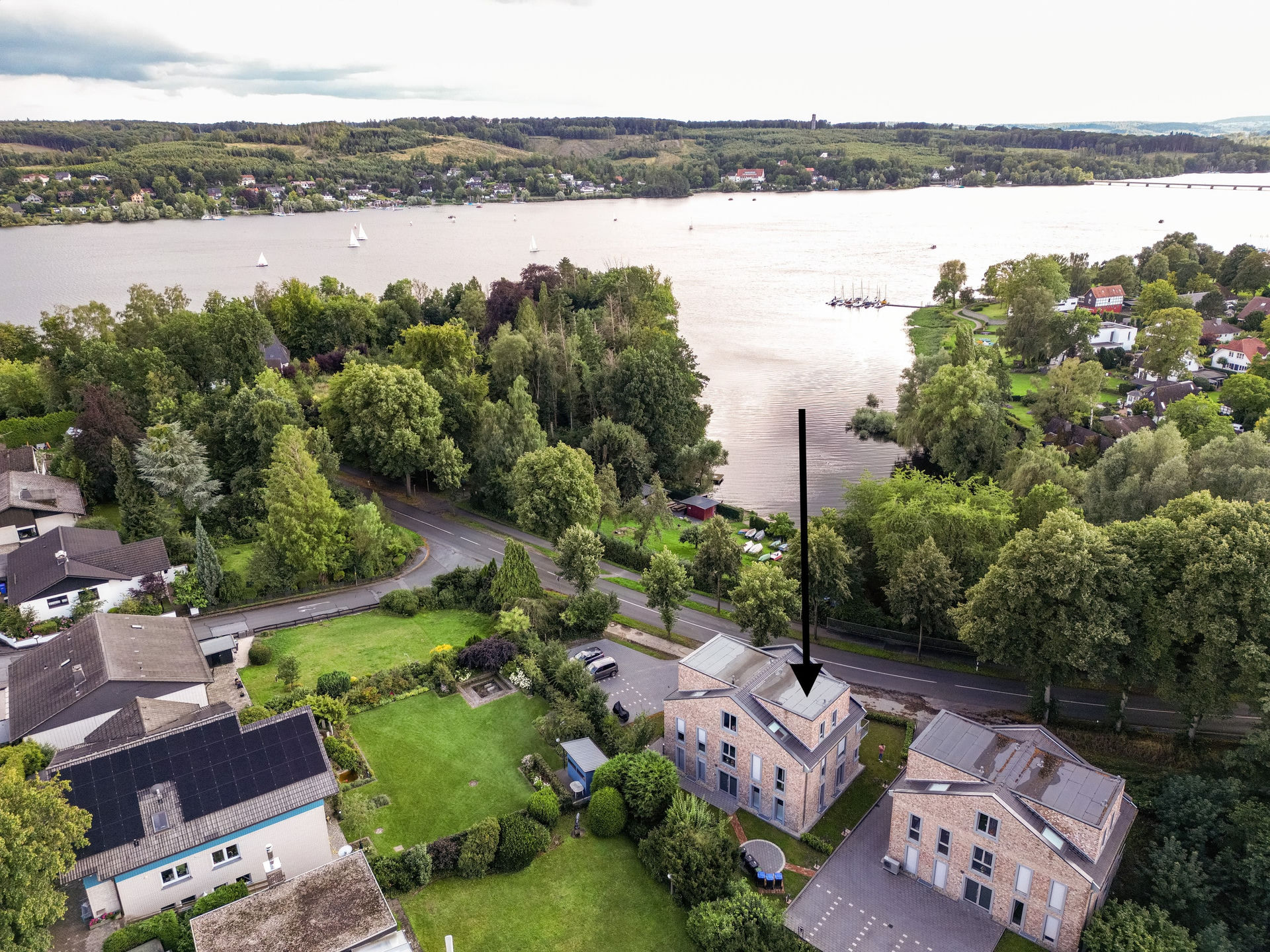 Aerial drone view of the brick houses and their lawns below a wide lake with sailing boats, with a black arrow overlay pointing at the roof of the building.